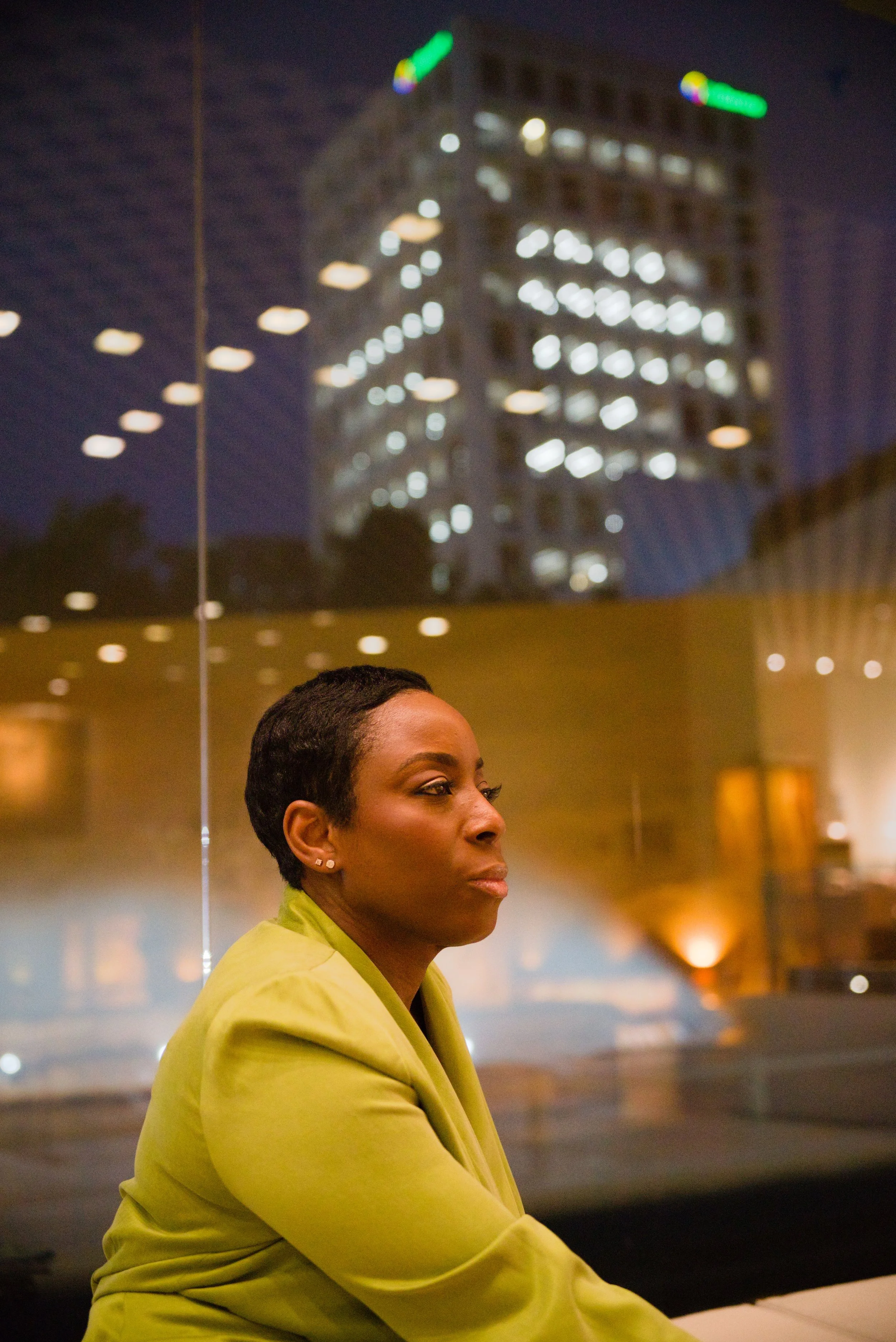 A woman with short dark hair and wearing a lime green blazer sitting in front of a glass window at night, with a high-rise building with illuminated windows behind her.