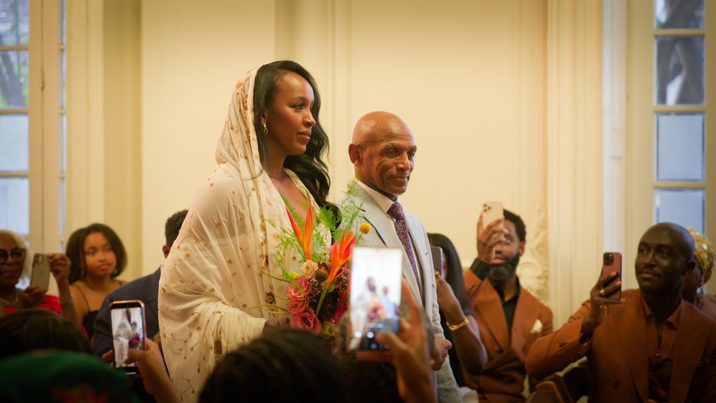 A woman in a cream-colored traditional dress holding a bouquet of flowers standing beside a man in a light gray suit at an indoor event, surrounded by people taking photos.