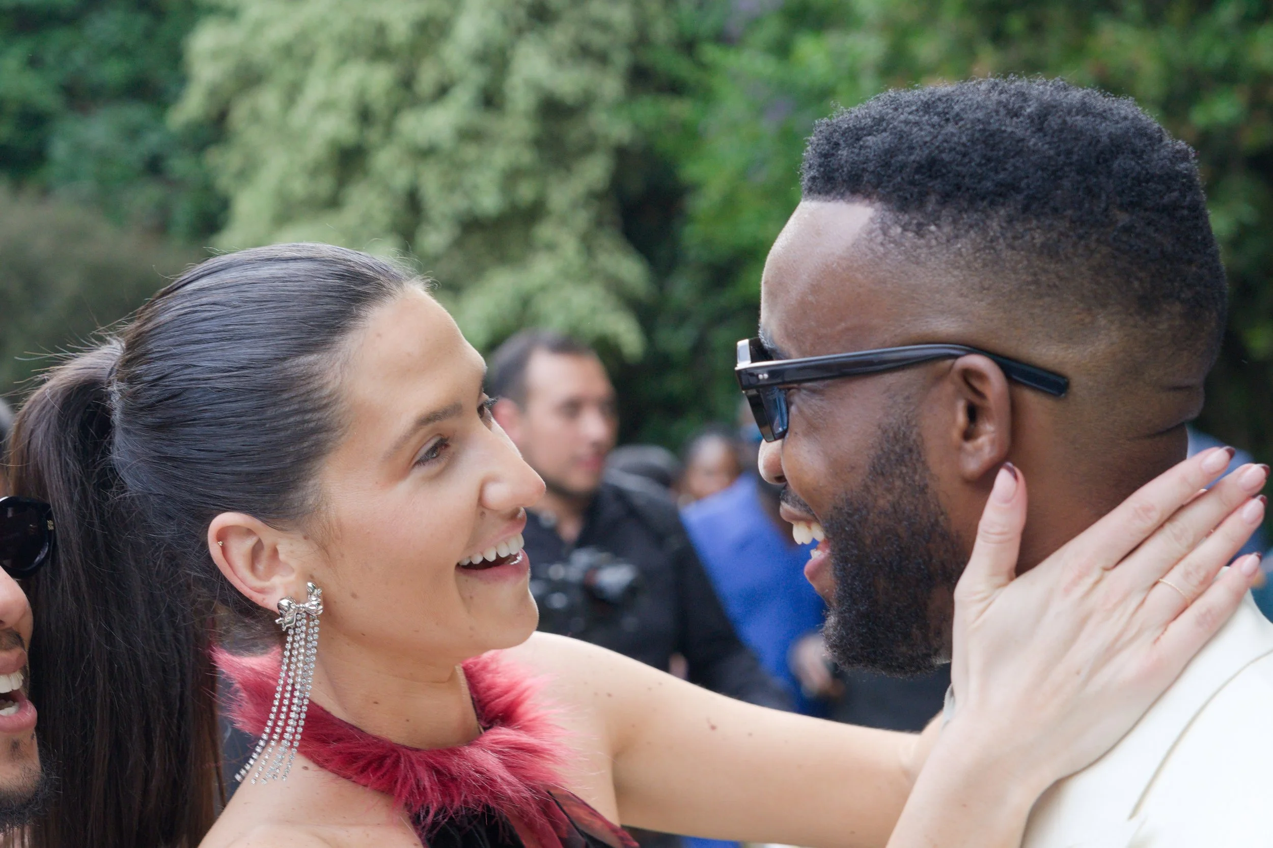 A woman and a man smiling and looking at each other, outdoors with greenery in the background. The woman has dark hair pulled back, earrings, and a dress with red feather detail. The man has short hair, a beard, glasses, and is wearing a white jacket