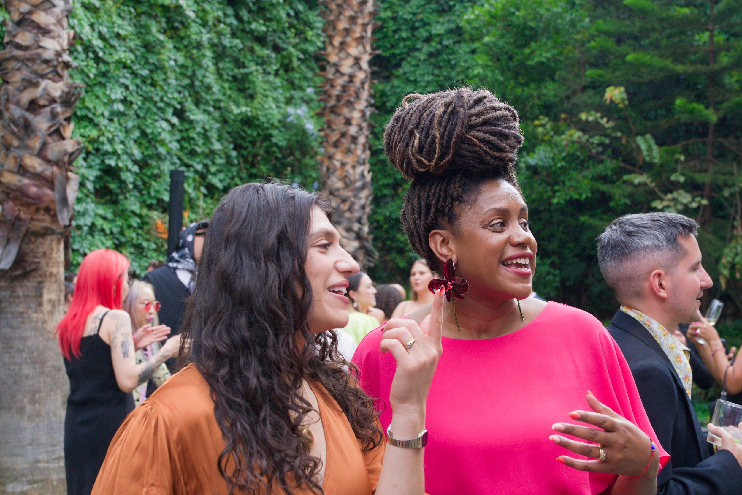 A group of people at an outdoor gathering, with two women in the foreground, one with dark wavy hair and the other with dark hair styled in large locs, smiling and chatting amidst lush green trees and other guests in the background.