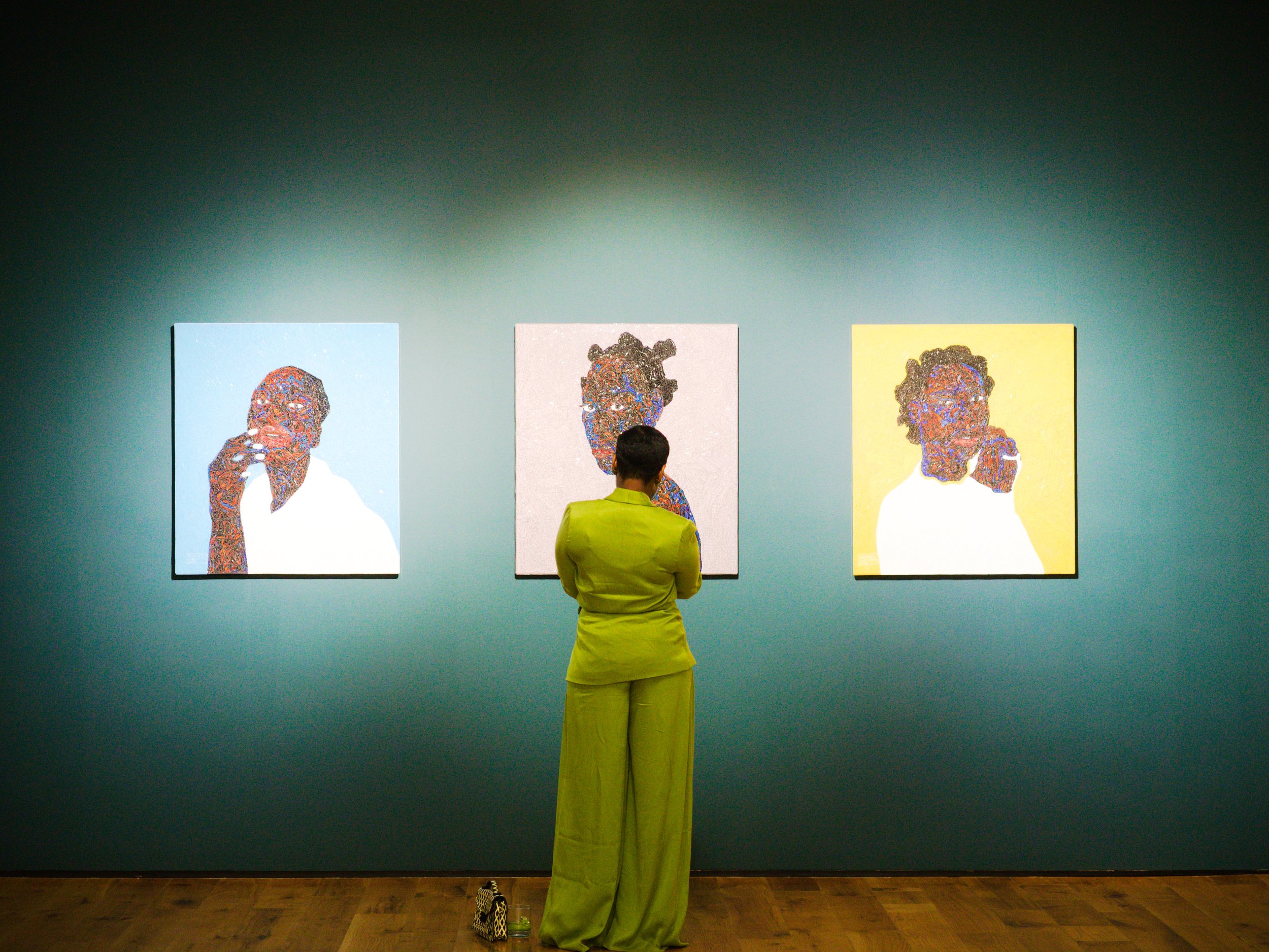 Person in lime green pants and top viewing three colorful portraits of women at an art gallery.