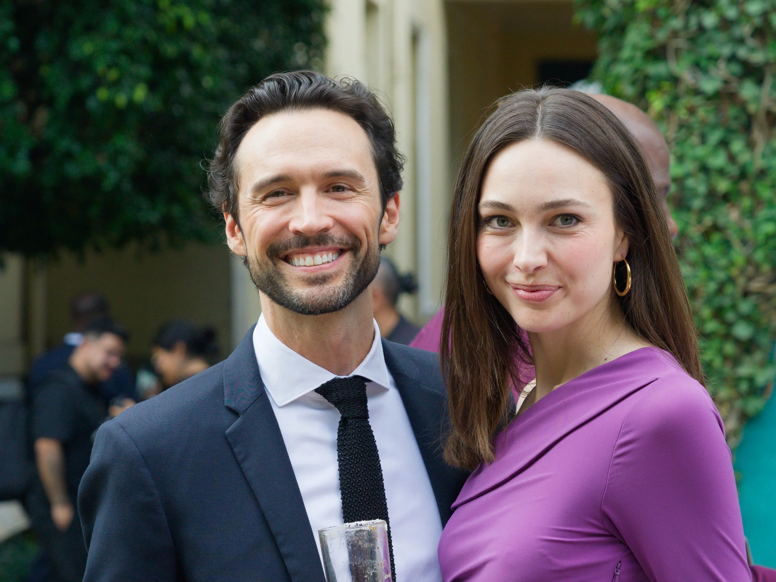 A man in a suit and a woman in a purple dress smiling at a social gathering outdoors.