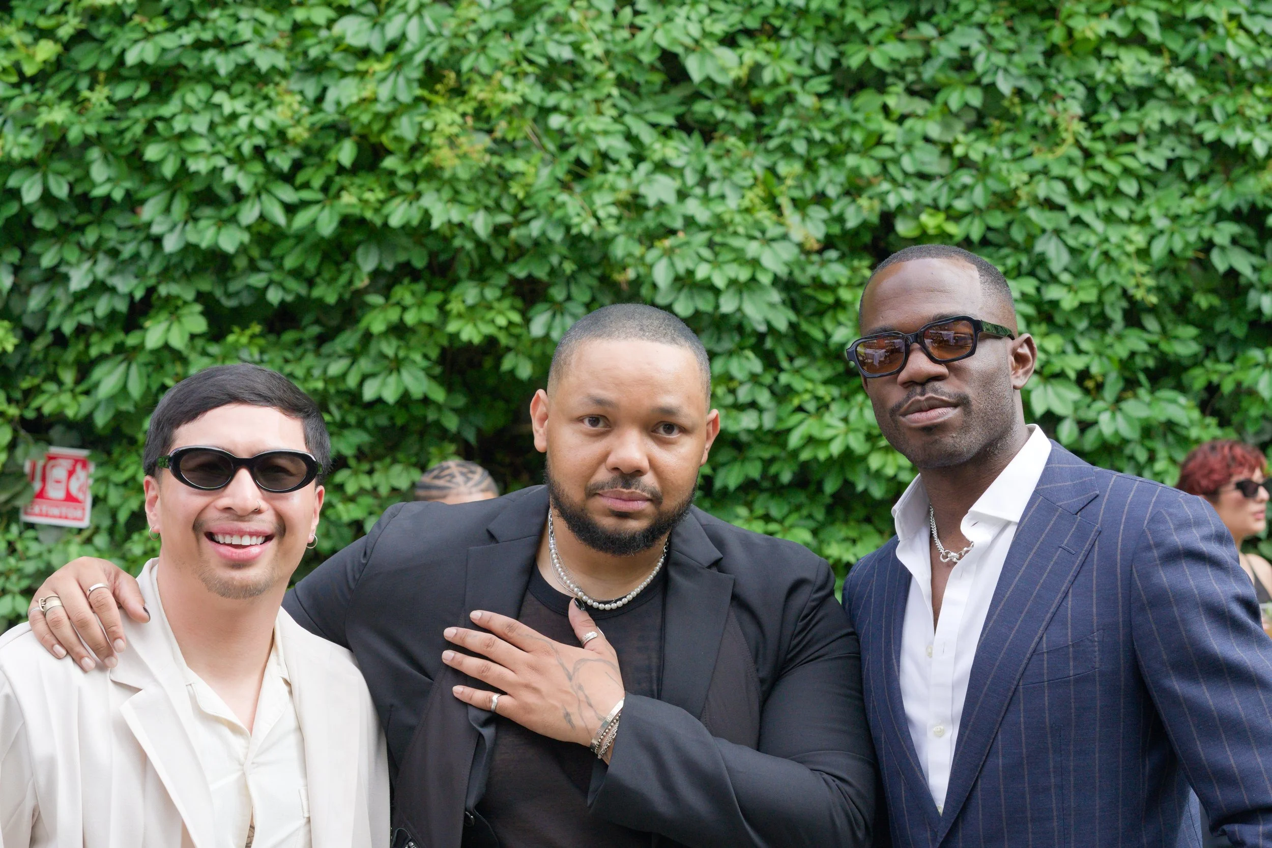 Three men standing together outdoors in front of green foliage, wearing stylish clothing and sunglasses, posing for the photo with friendly expressions.