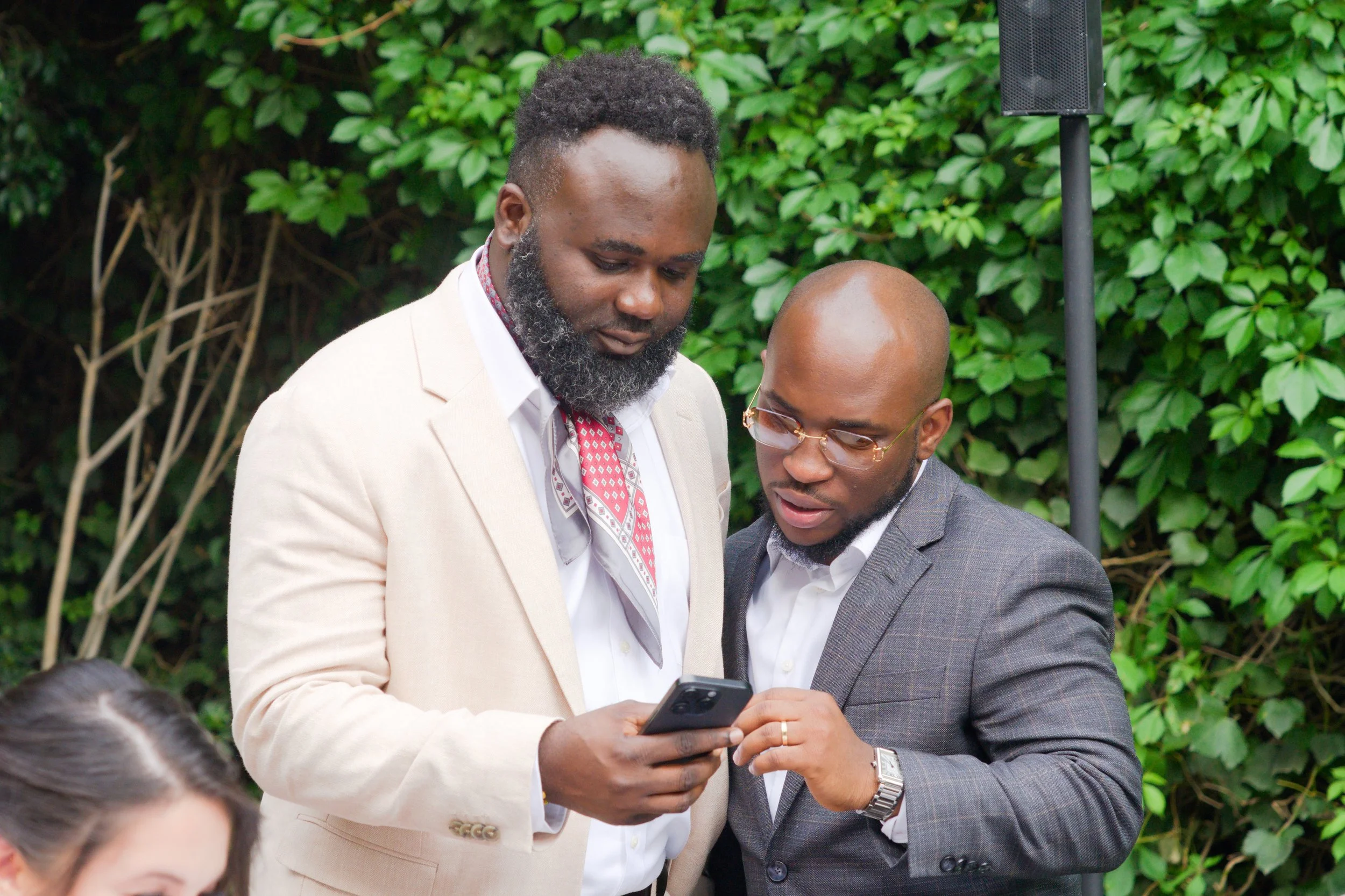Two men in suits looking at a smartphone together outdoors with green foliage in the background.