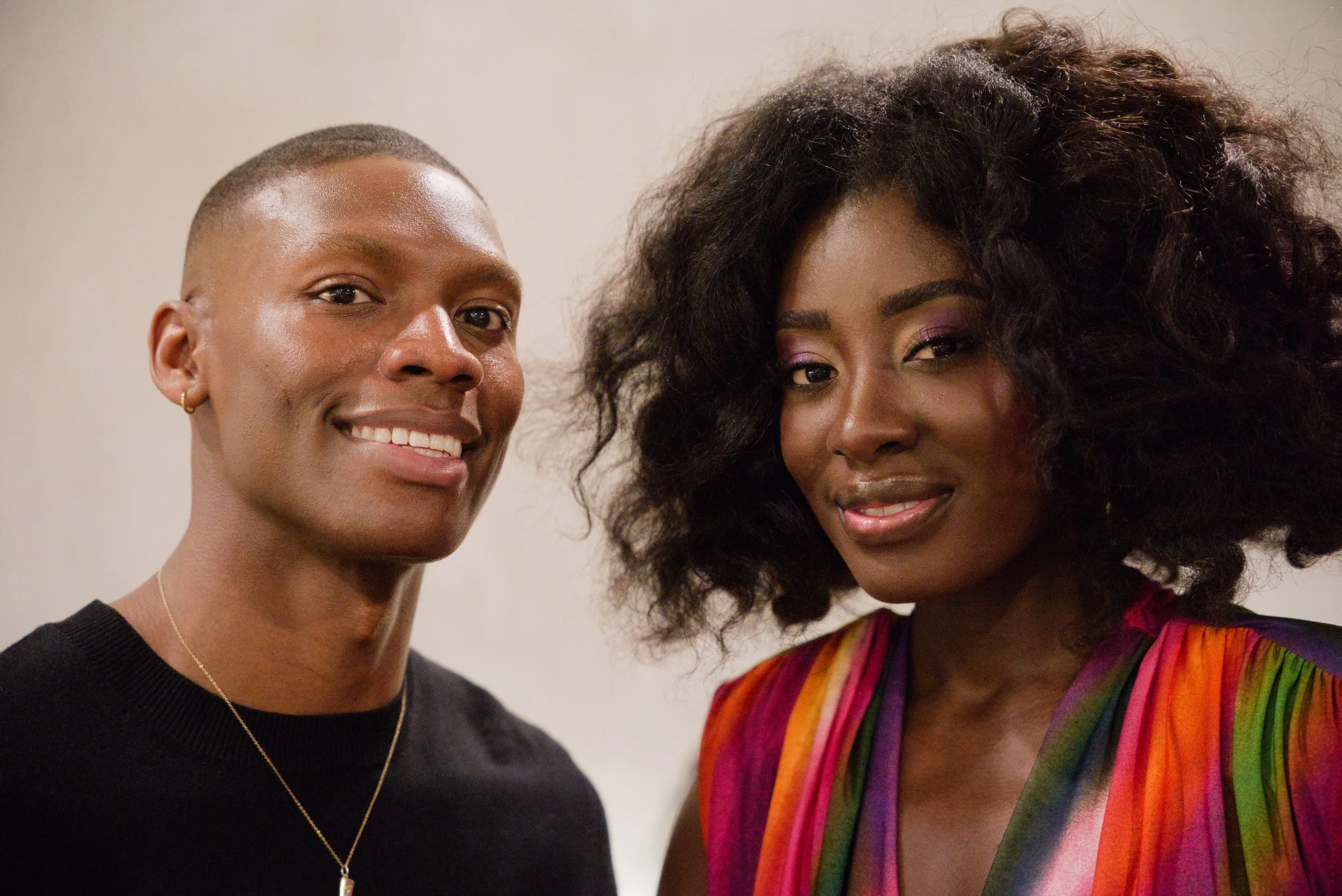 A smiling man with short hair, wearing a black shirt and a necklace, standing next to a woman with voluminous curly hair, wearing a colorful rainbow-patterned dress, both looking at the camera.