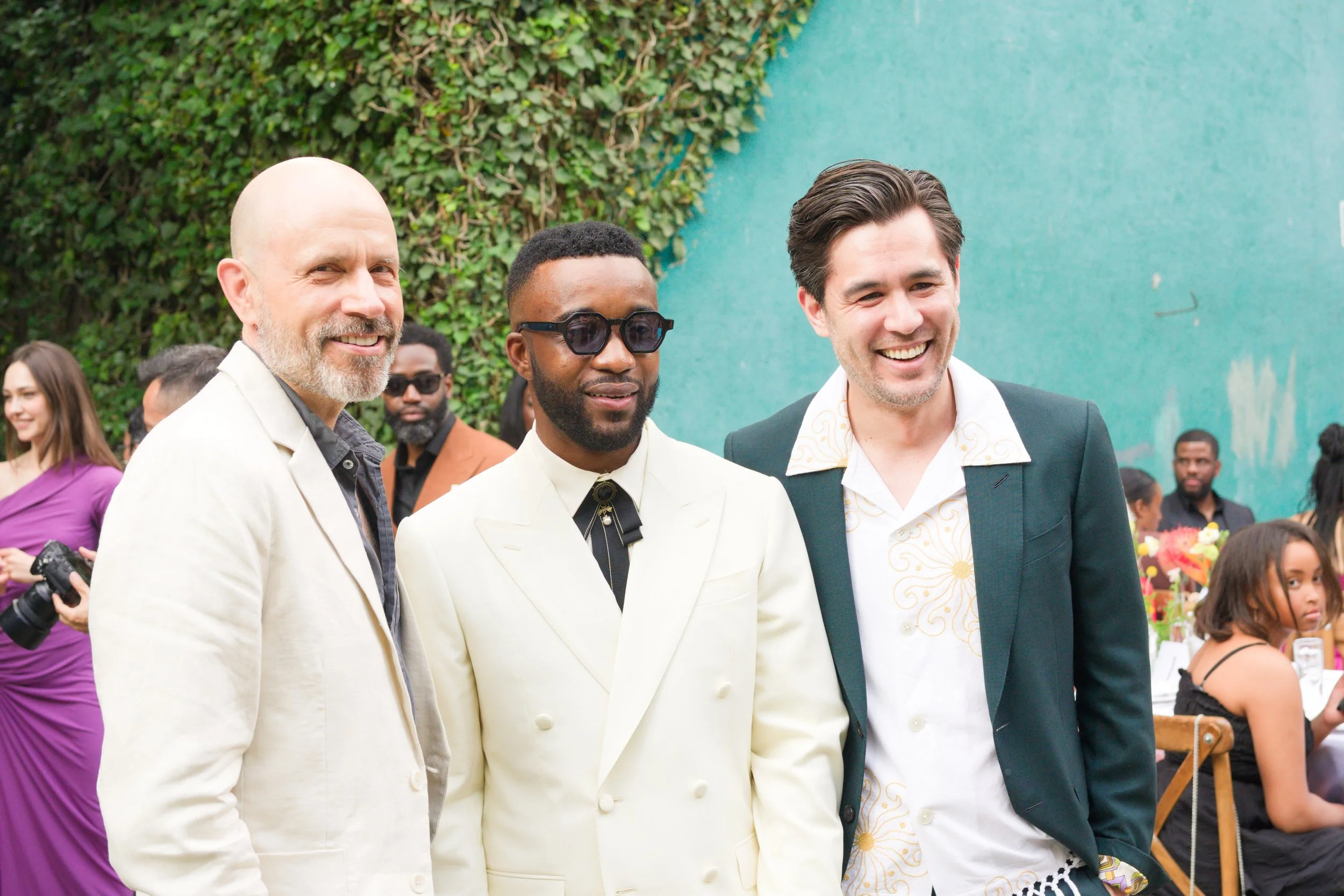 Group of people smiling at an outdoor event, with three men in the foreground and others in the background, some holding cameras, with greenery and a teal wall behind them.