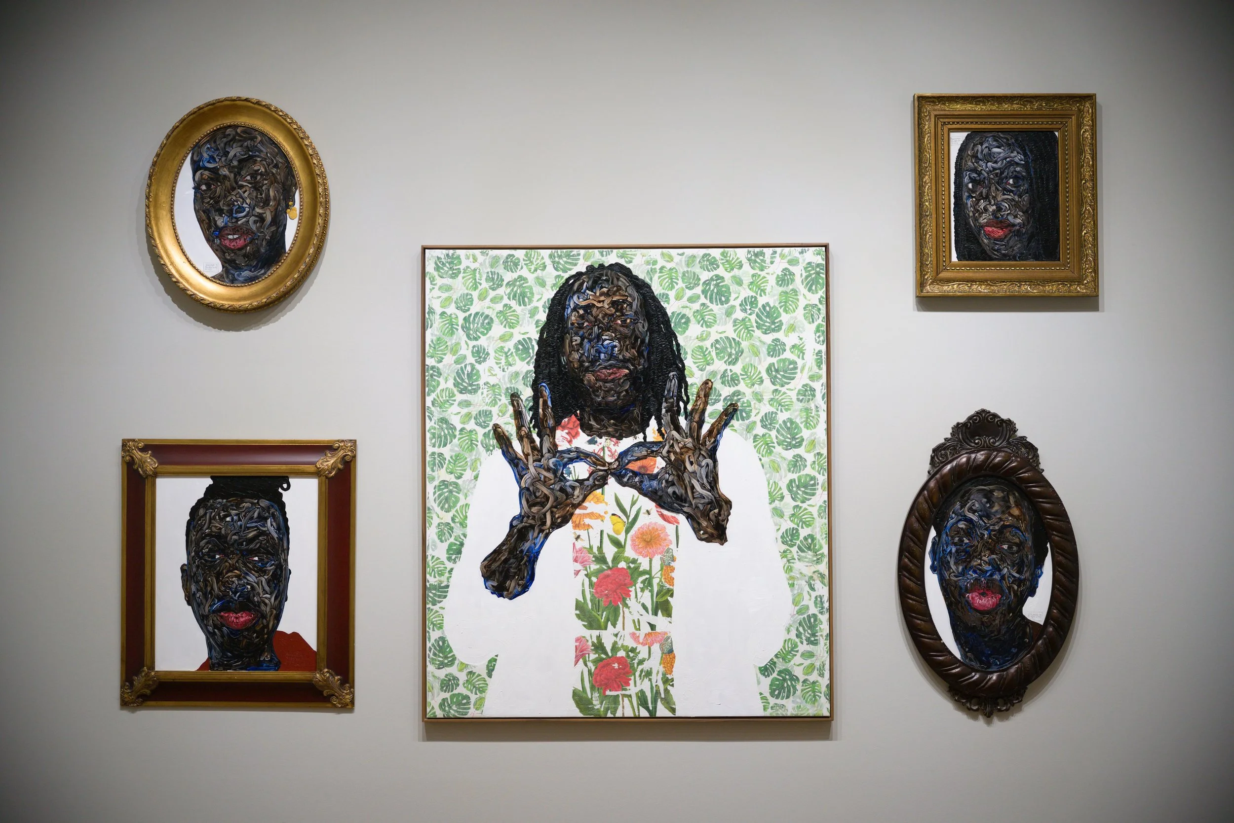 Art gallery wall with five framed portraits featuring a woman with dark skin, curly hair, and red lips, created with layered, textured materials. The central piece depicts the woman with floral and leafy backgrounds, and her hand forming a peace sign