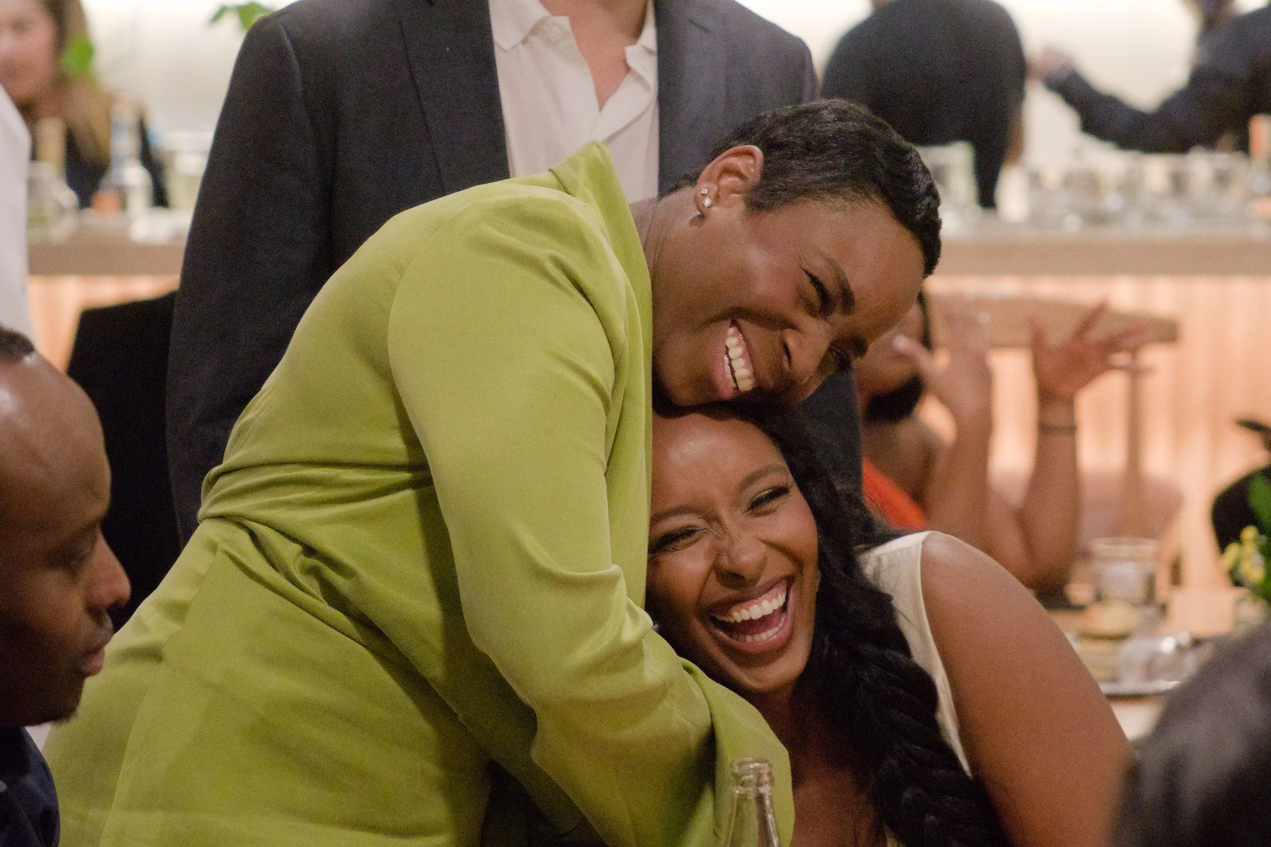 Two women and a man at a social gathering, with the women laughing and embracing each other, and the man standing behind them, smiling broadly.