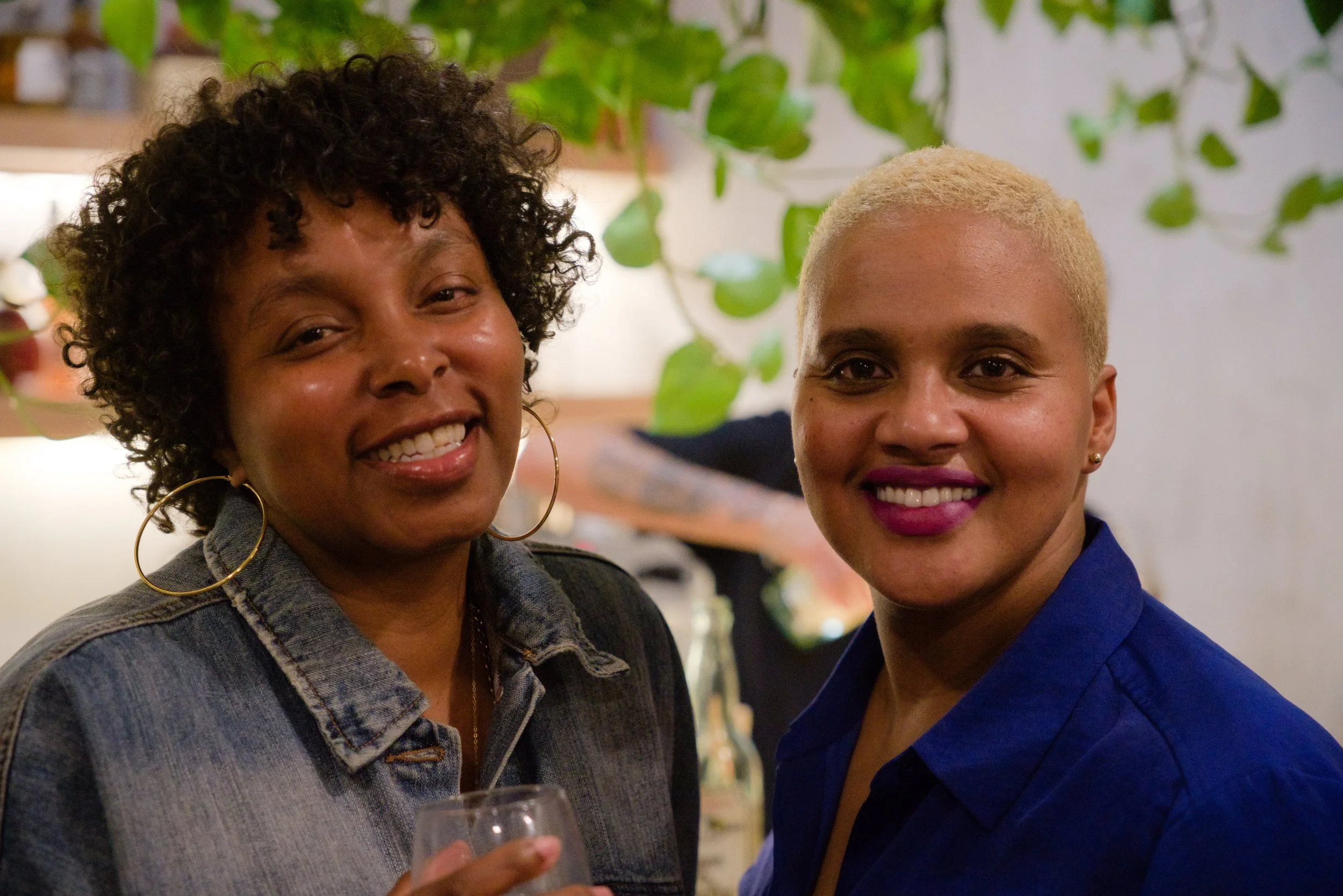 Two women smiling at a social gathering indoors, with green plants and decorated background, one holding a glass.
