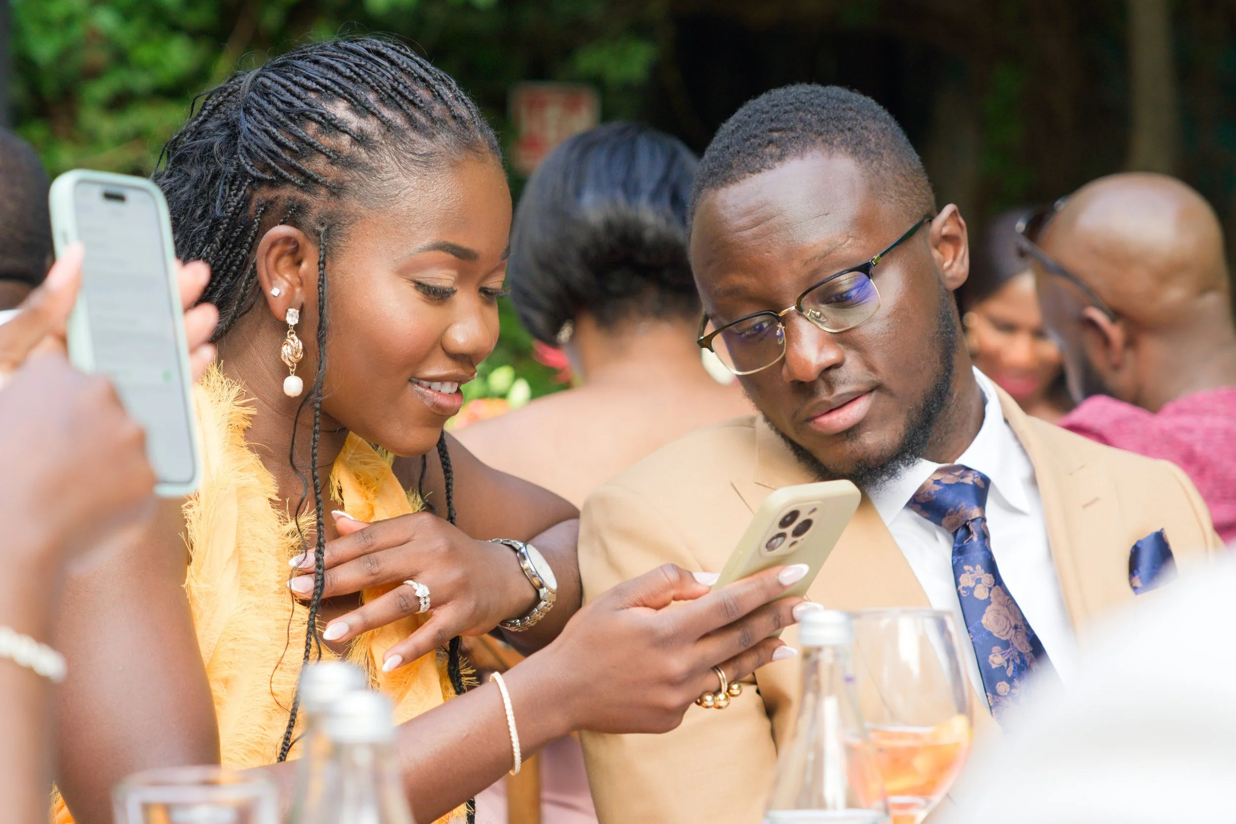 Two people, a woman and a man, looking at a smartphone together at a social event outdoors. The woman has braided hair and is wearing earrings and a yellow dress. The man wears glasses, a tan suit, and a blue tie. Both appear engaged and smiling at t