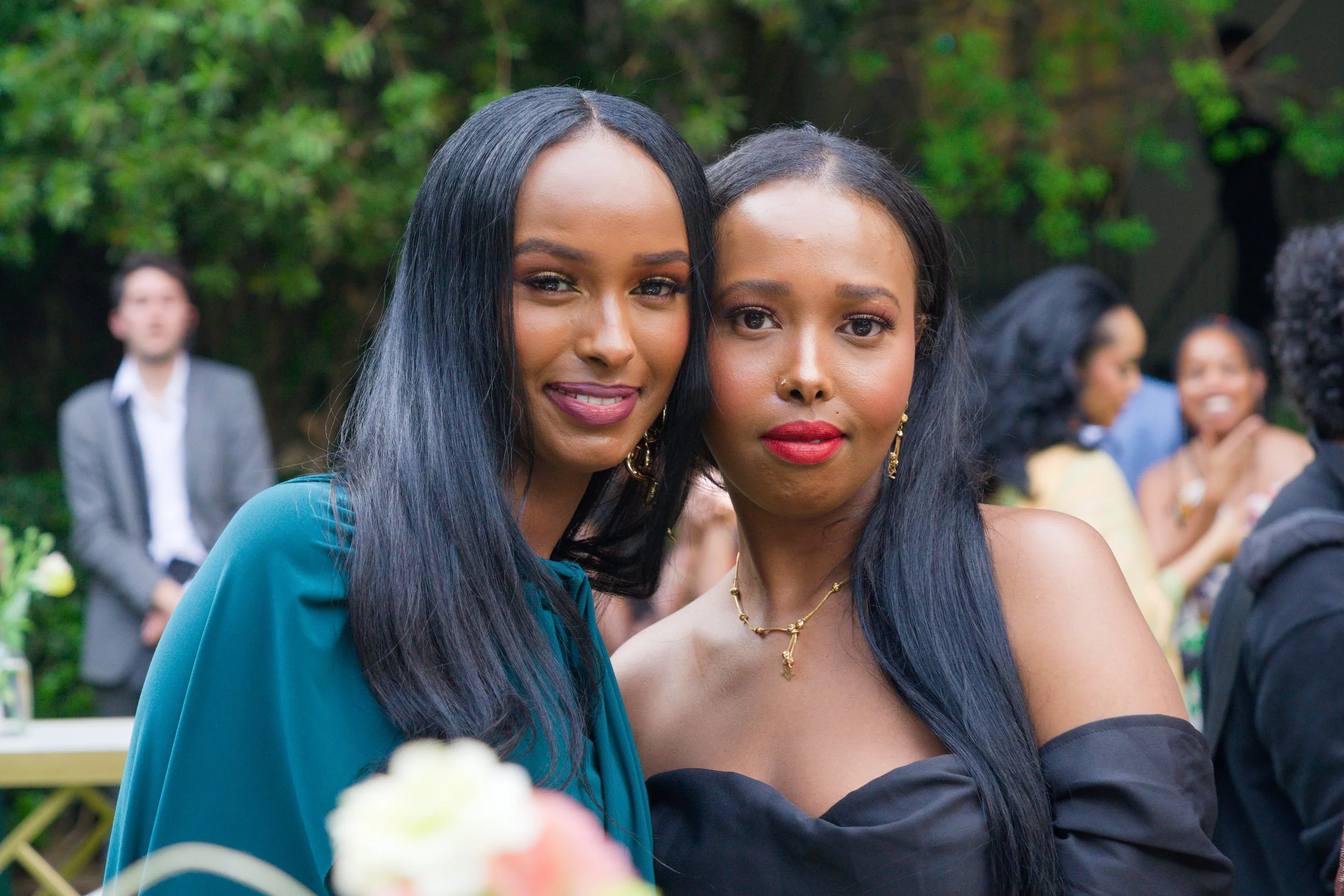 Two women with long black hair dressed formally, posing outdoors during a social event with other people in the background.