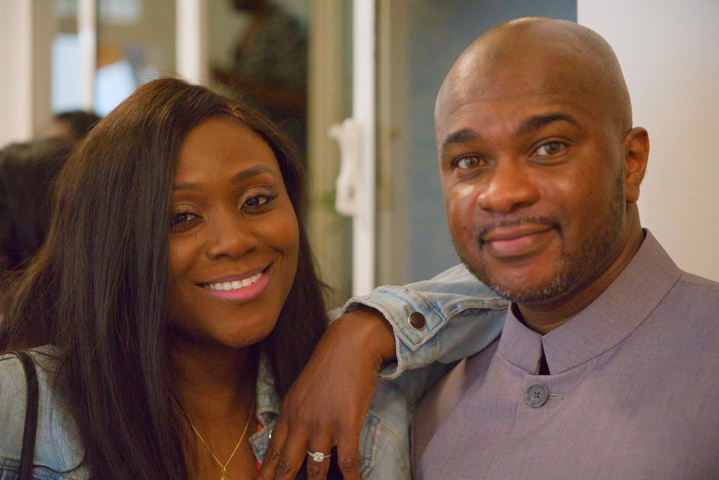 A smiling woman with dark hair and a man with a shaved head and beard, standing close together indoors, with the woman resting her arm on the man's shoulder.