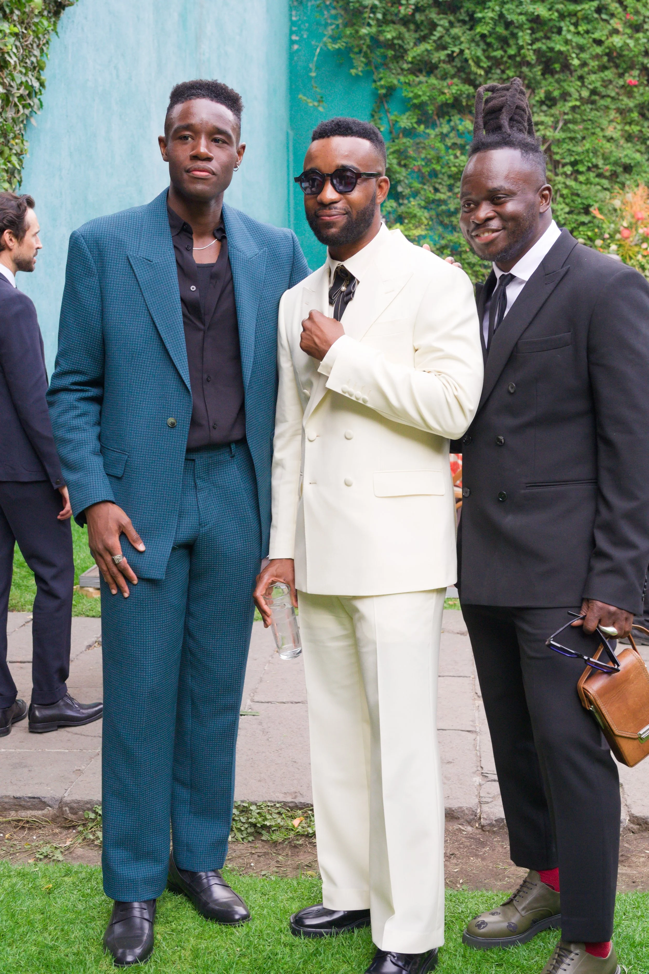 Three men in suits standing outdoors, smiling, with a green and blue wall and foliage in the background.