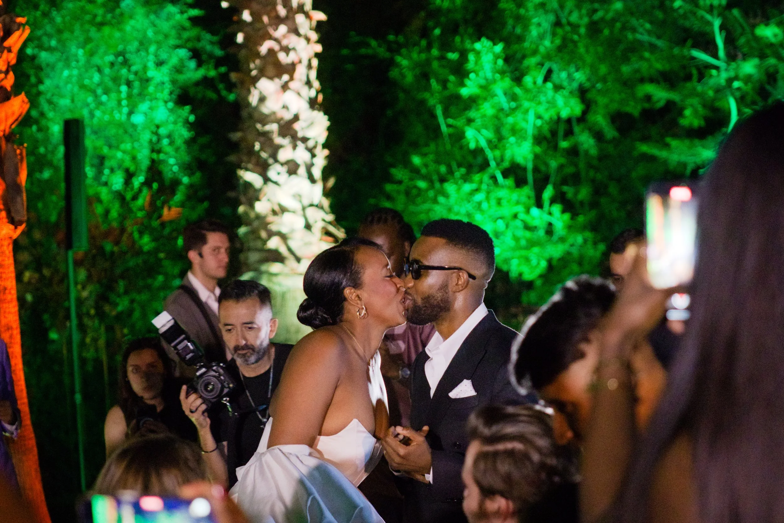 Couple sharing a kiss at a wedding reception, illuminated by green and white lights, surrounded by guests and a photographer.