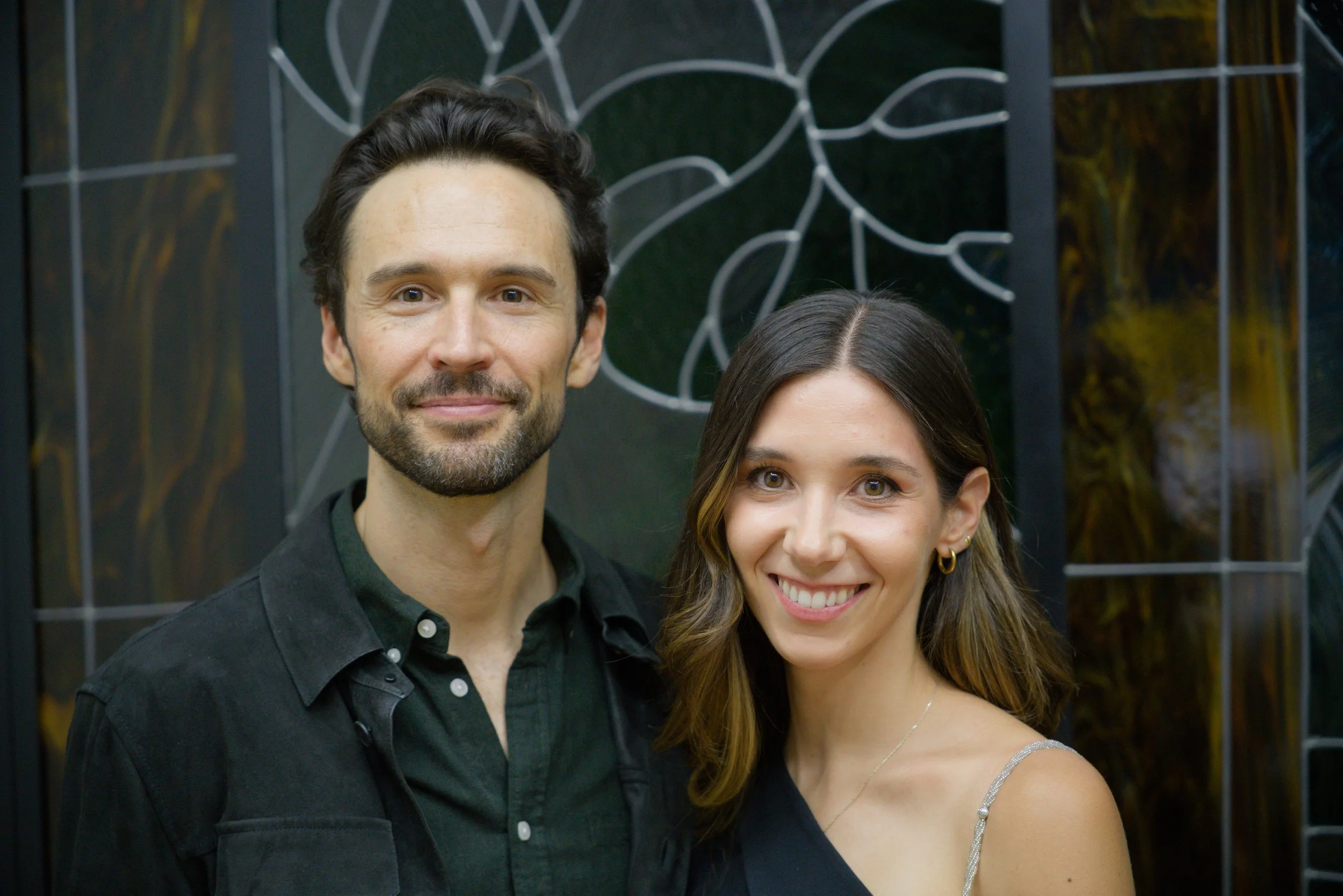 A man and a woman smiling, standing close together in front of an artistic wall design.