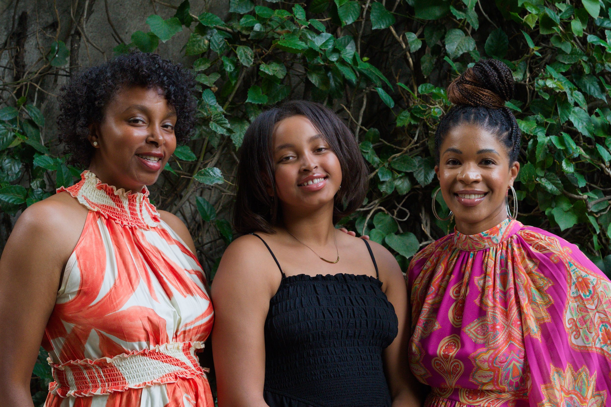 Three women standing together outdoors in front of green leafy background, smiling at the camera.