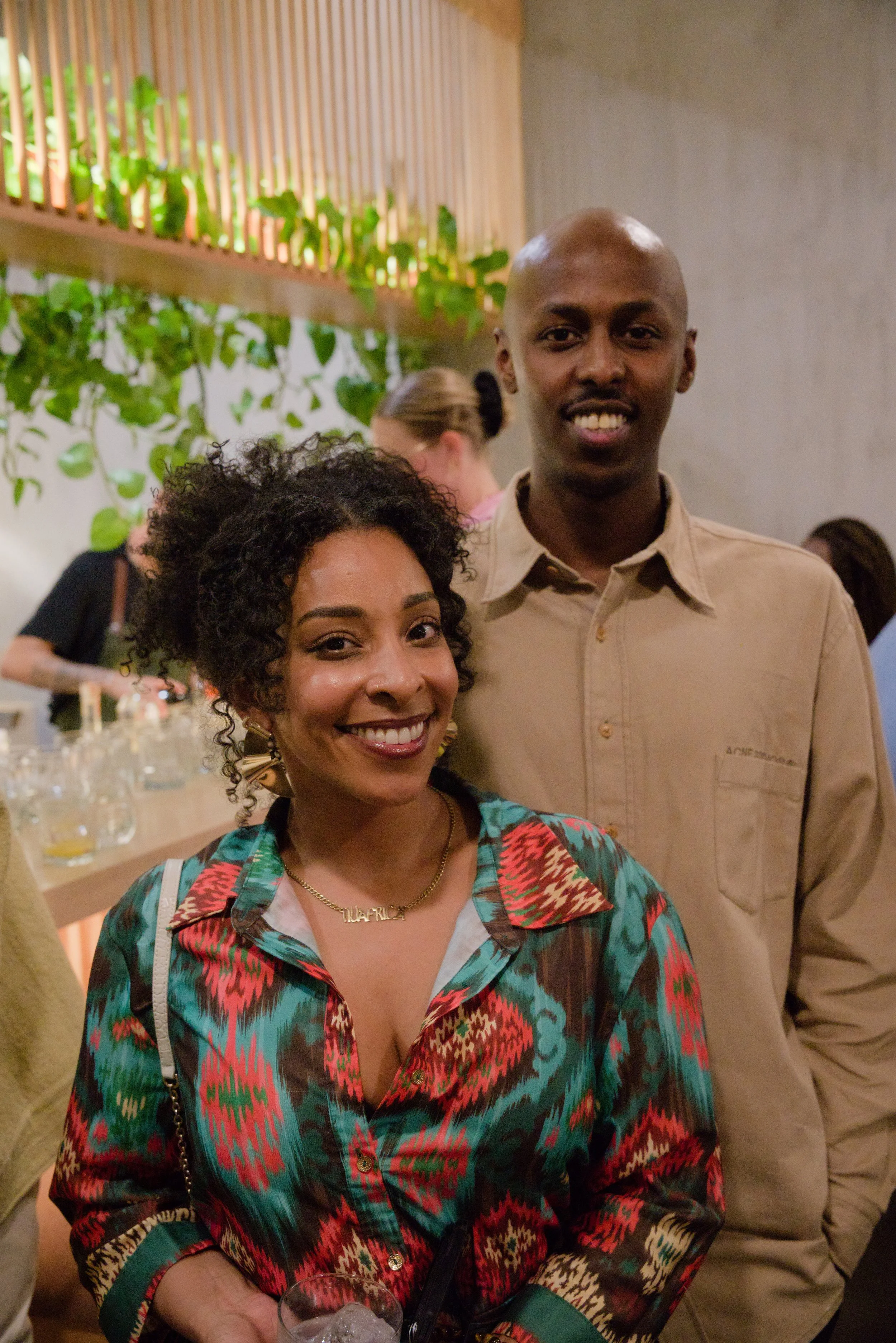 A smiling woman with curly hair and a man with a bald head pose together at a social event, with a bar and other people in the background.
