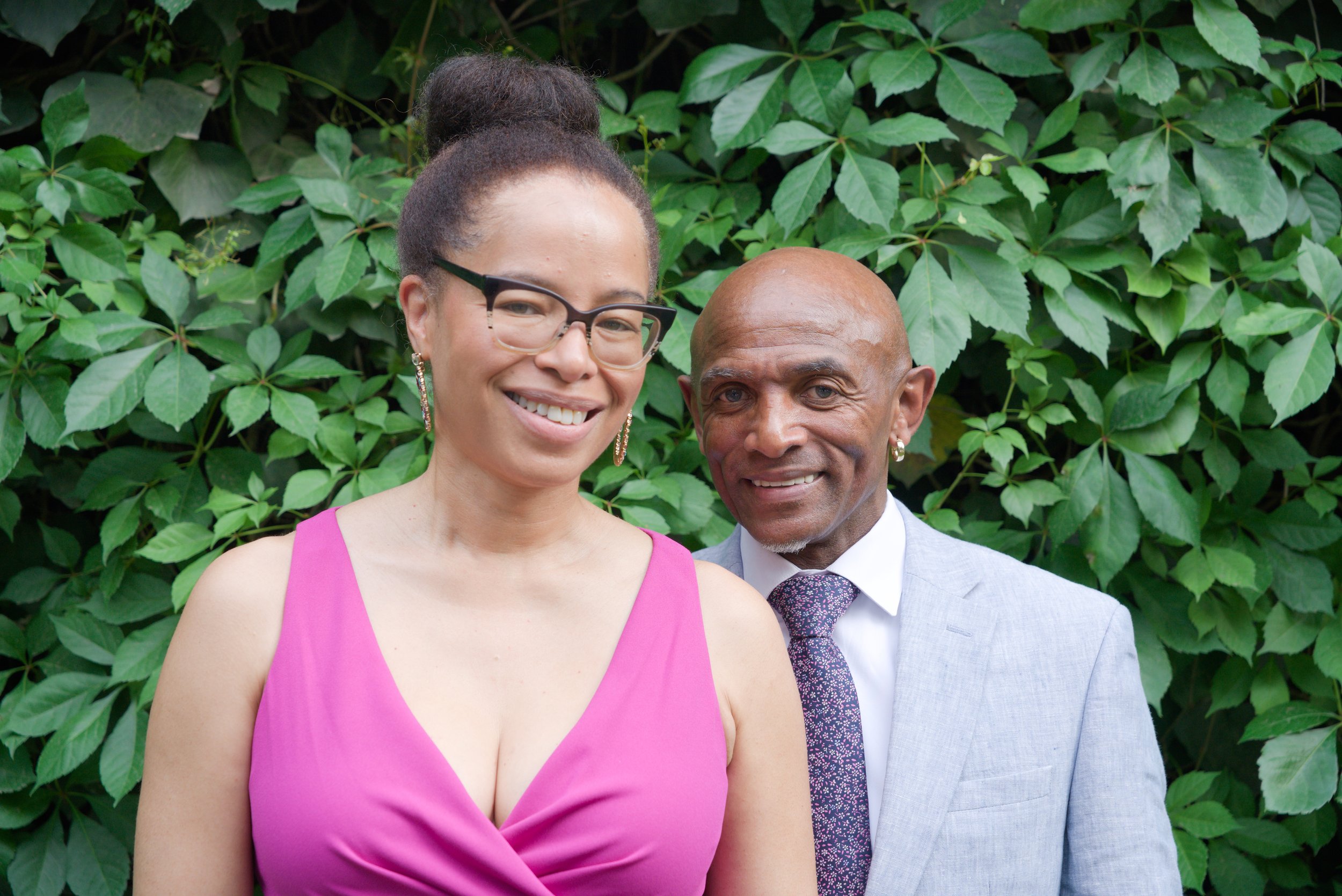 A smiling woman in a pink dress with glasses and earrings stands next to a smiling man in a light gray suit with a white shirt, purple tie, and earrings. They are outdoors against a backdrop of green leaves.
