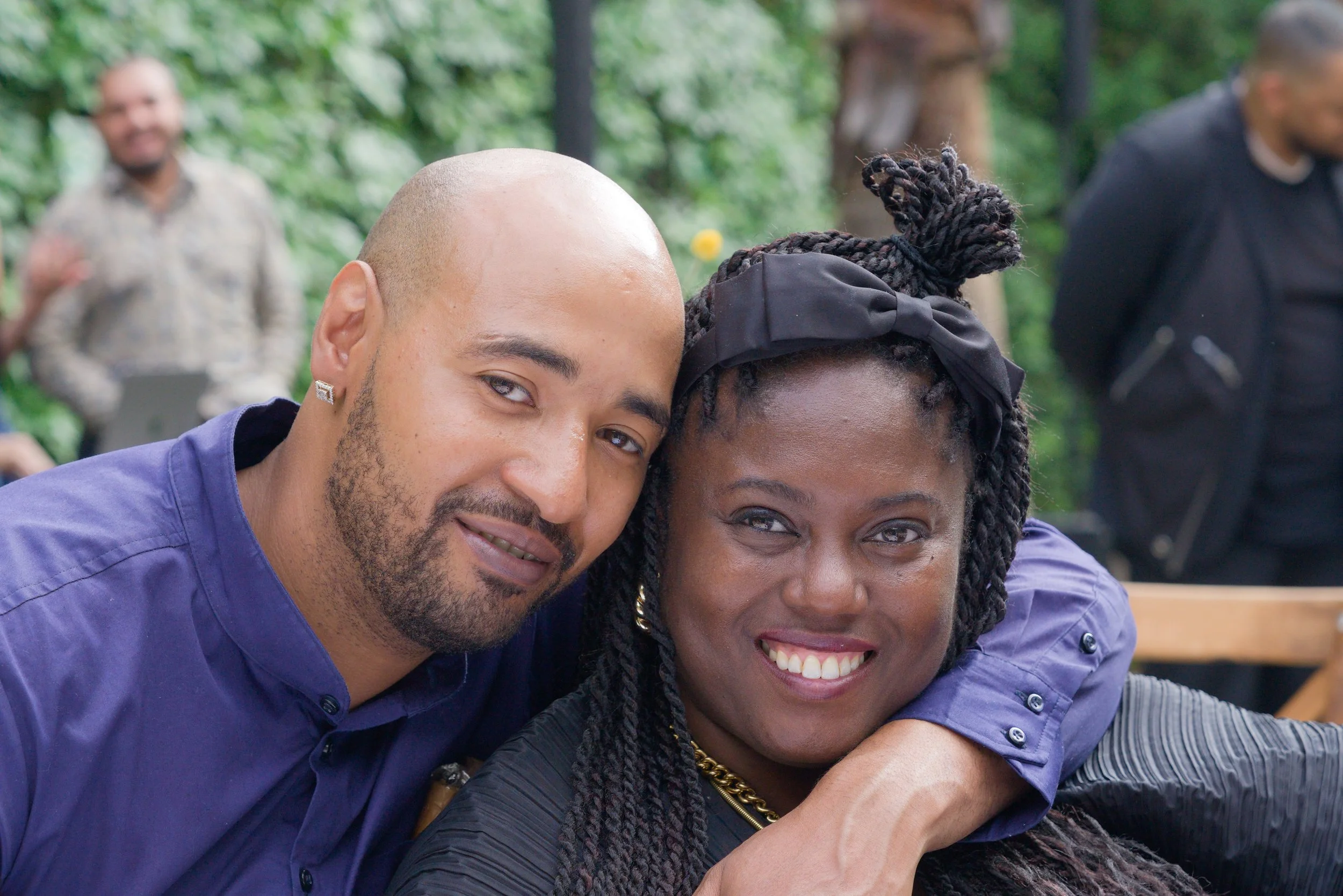 A close-up of a smiling man and woman embracing each other outdoors with a blurred background of people and greenery.