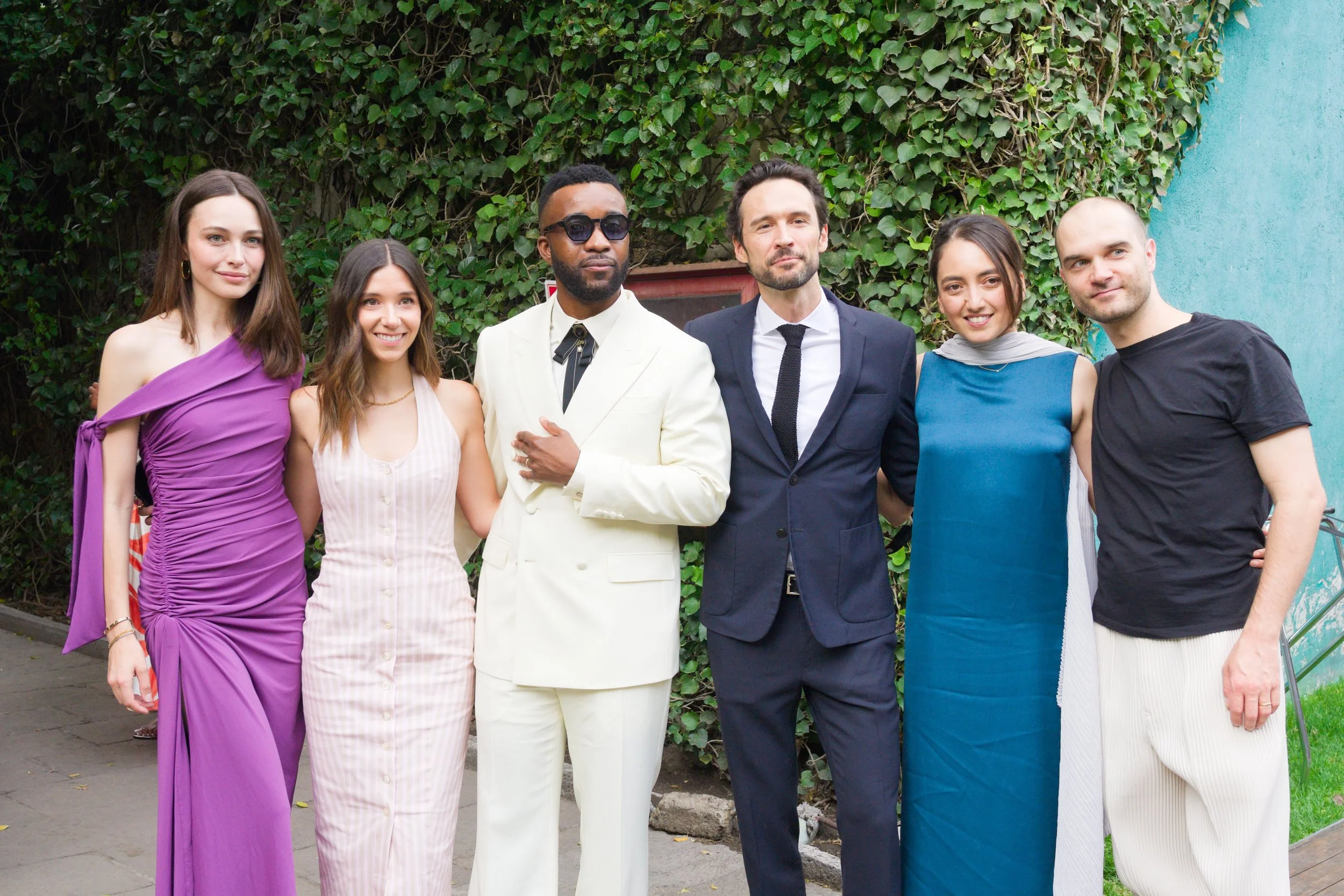 Group of six diverse people standing outdoors in front of green vine-covered wall, dressed in formal and semi-formal attire, smiling and posing for the photo.