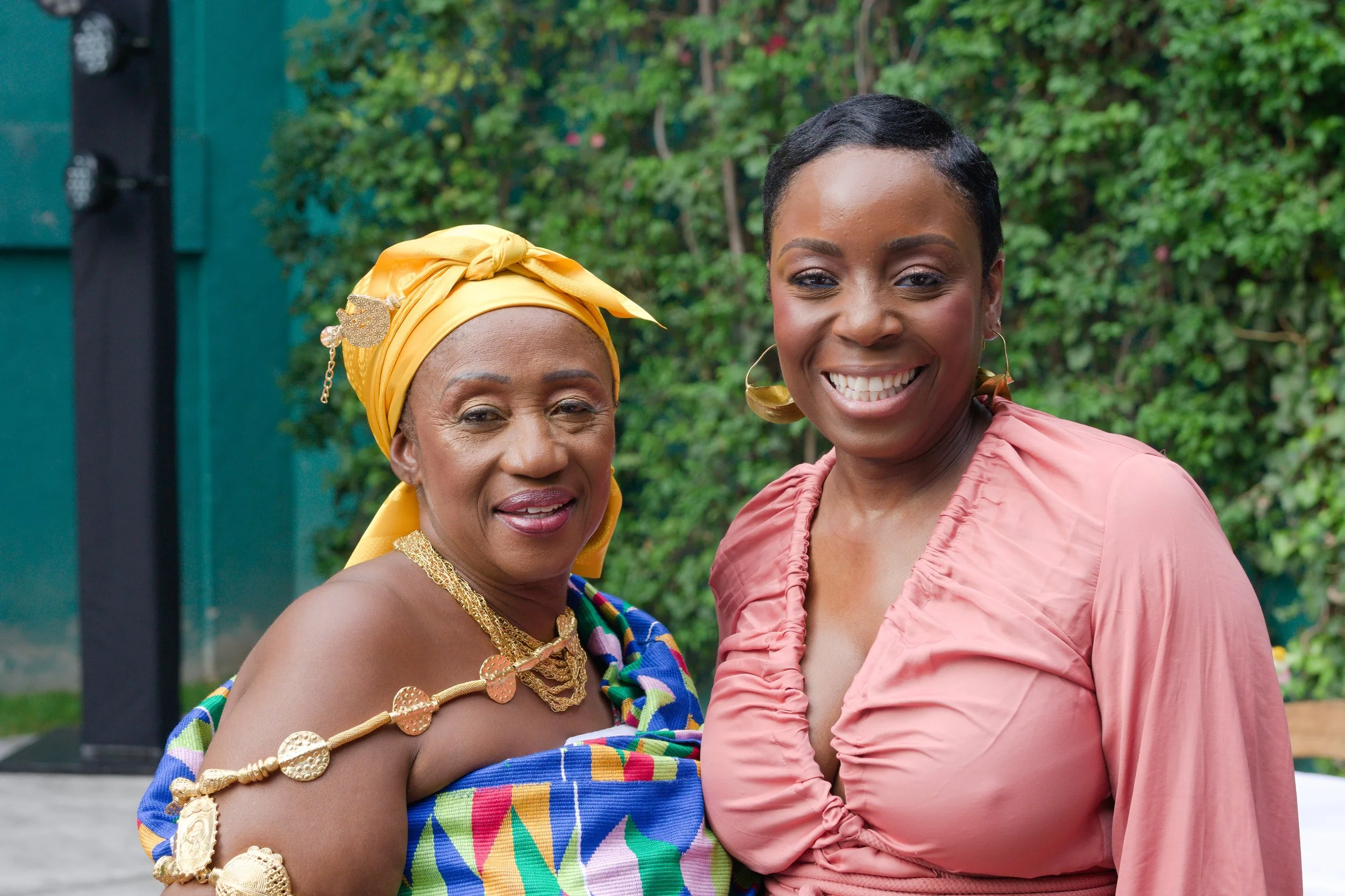 Two women smiling outdoors, one wearing vibrant traditional African attire with gold jewelry and a yellow headwrap, the other in a pink dress with earrings, standing in front of green foliage.