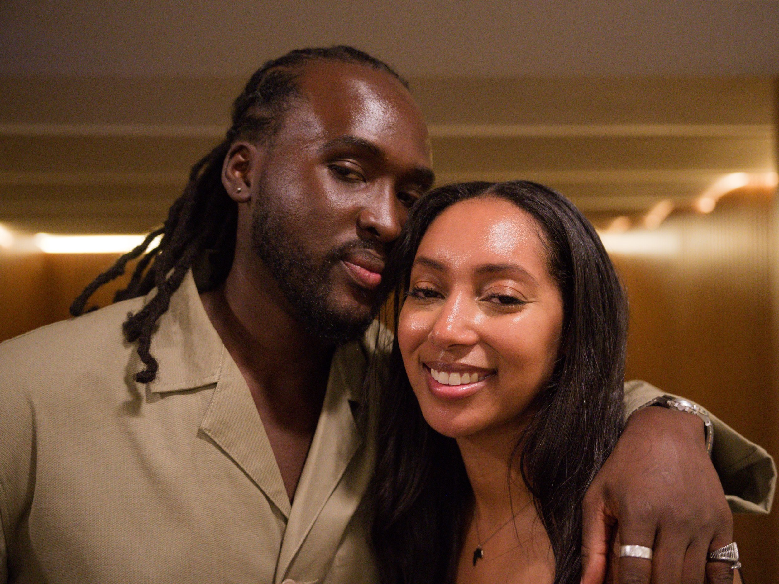 A man and woman smiling closely, with the man's arm around the woman's shoulder, indoors.
