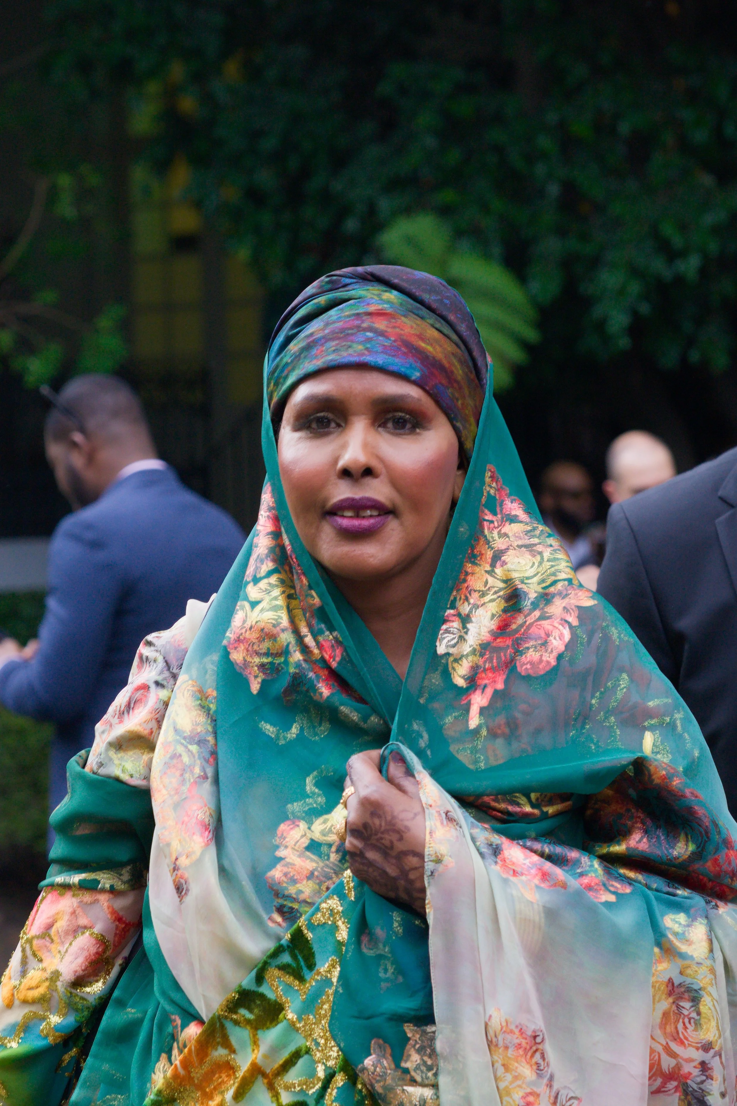 A woman wearing a colorful head covering and floral shawl, standing outdoors among other people.