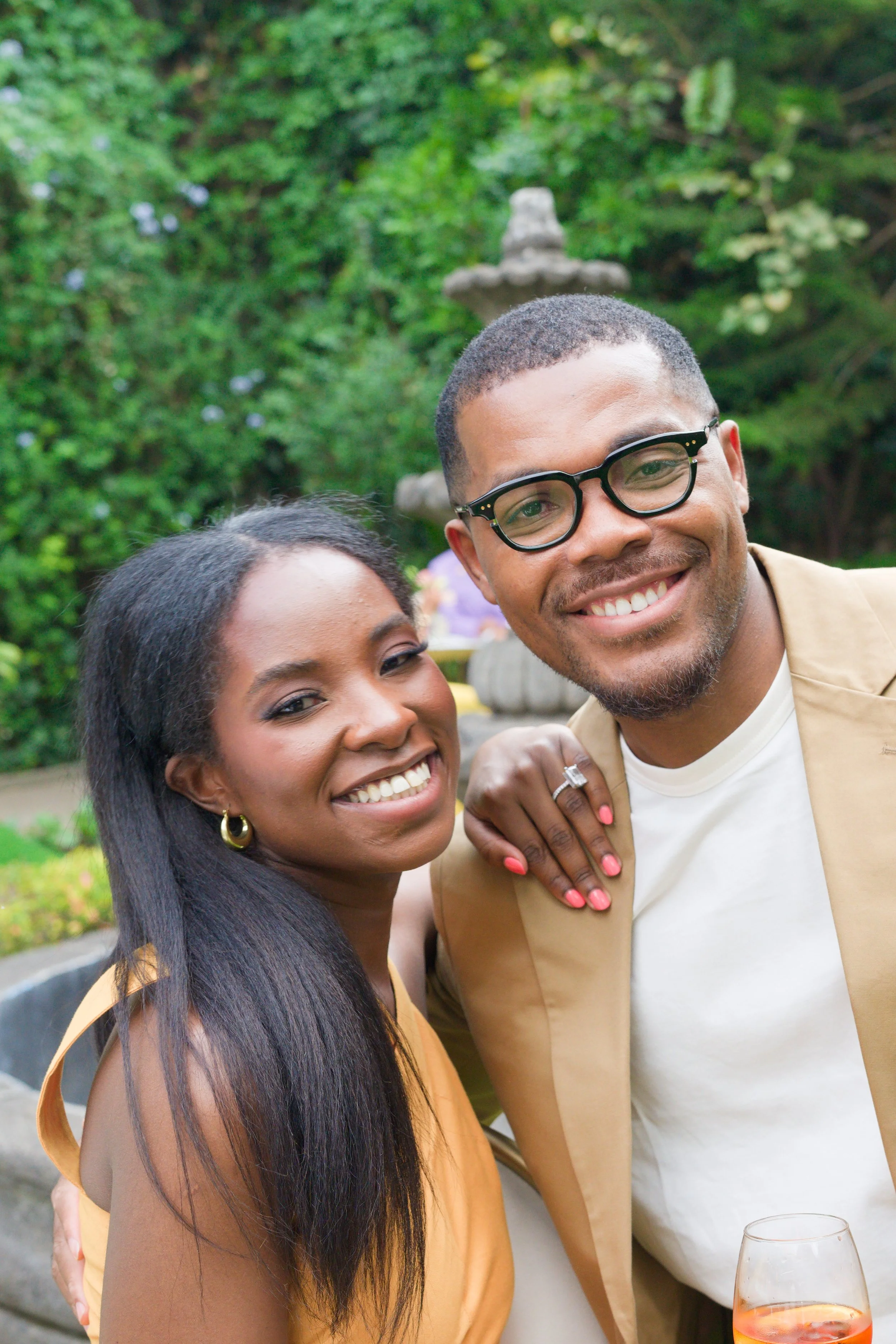 A smiling man and woman at an outdoor gathering, with greenery and a stone fountain in the background. The woman has dark hair and is wearing yellow clothing with gold earrings. The man has short hair, glasses, and is wearing a tan blazer and white t