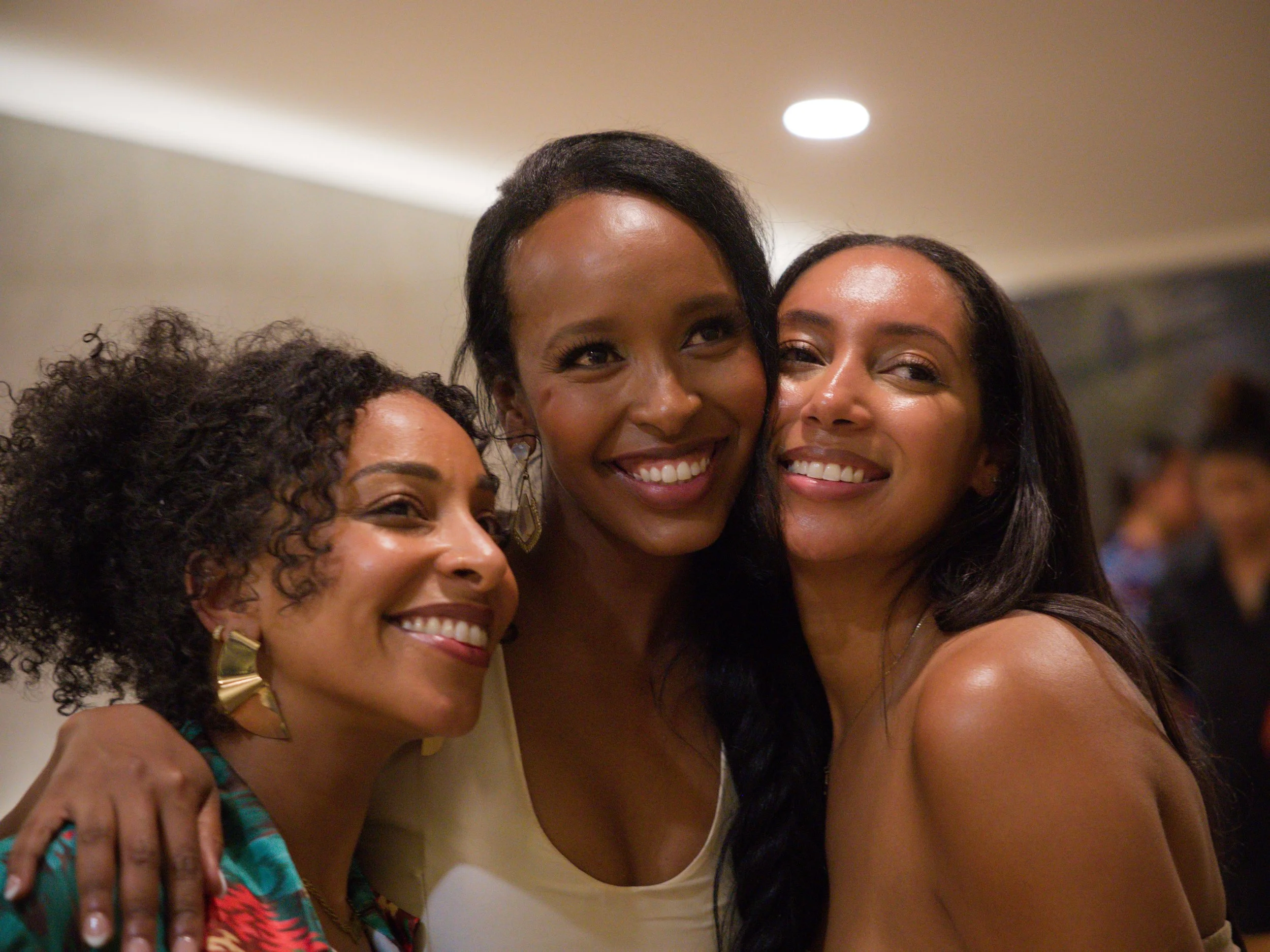 Three women smiling and hugging each other at an indoor event.