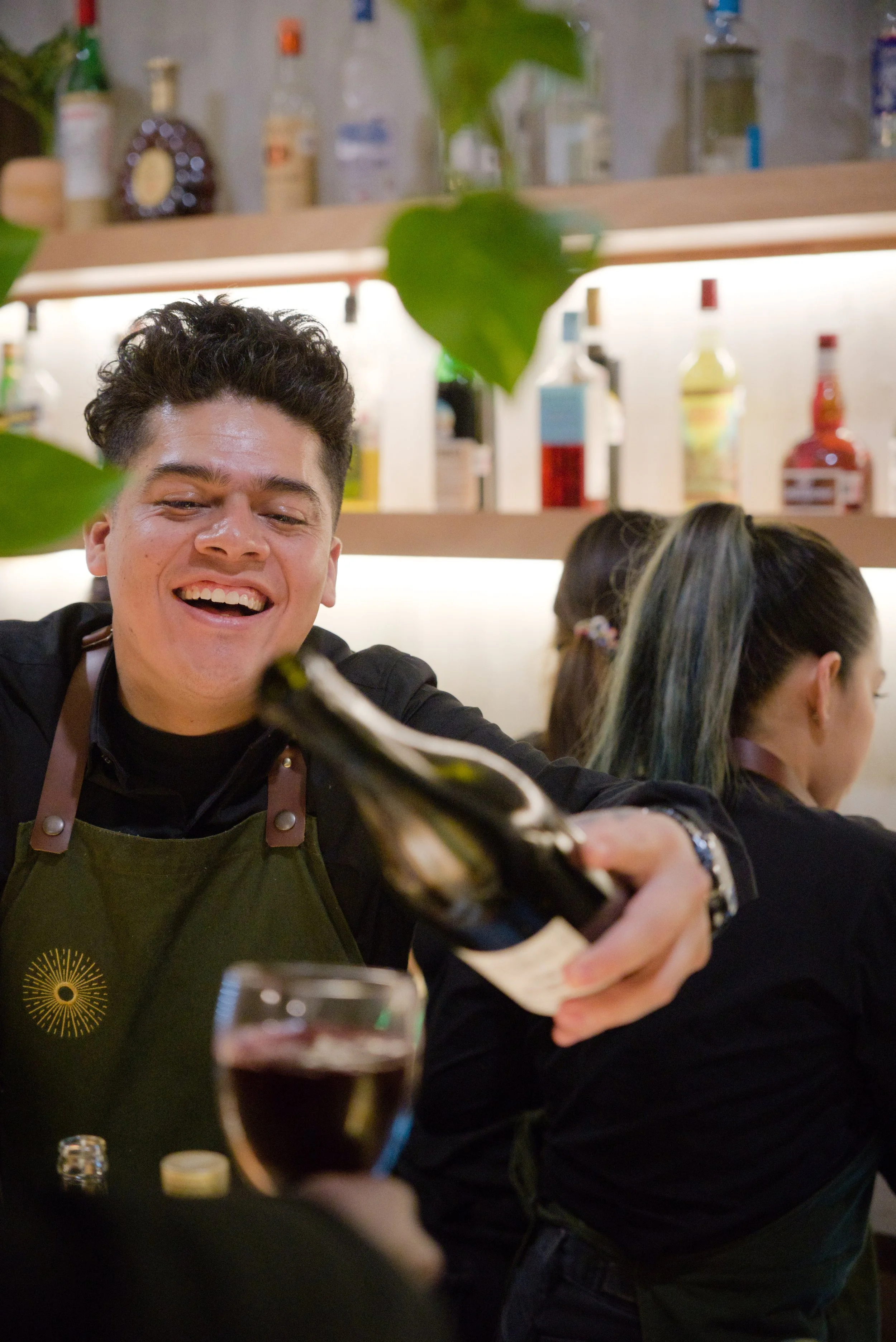A smiling bartender pouring a drink at a bar, with bottles on shelves behind him, and two women sitting at the bar in the background.