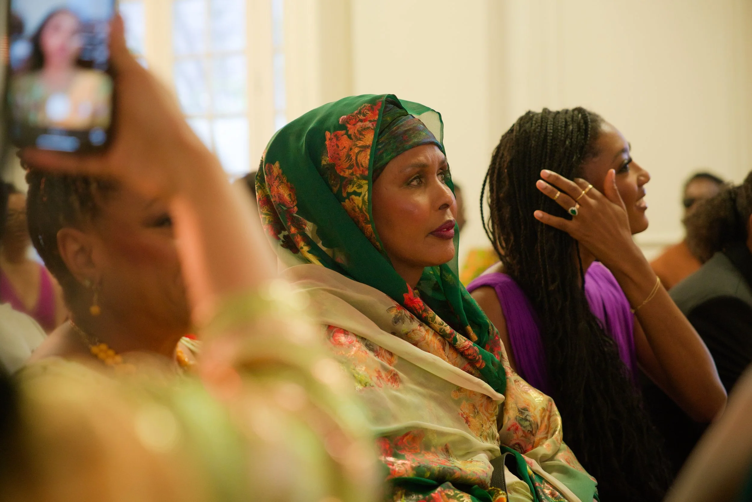Women attending an indoor event, focusing on a woman wearing a green and floral headscarf, sitting next to a woman in a purple top, with other attendees in the background.