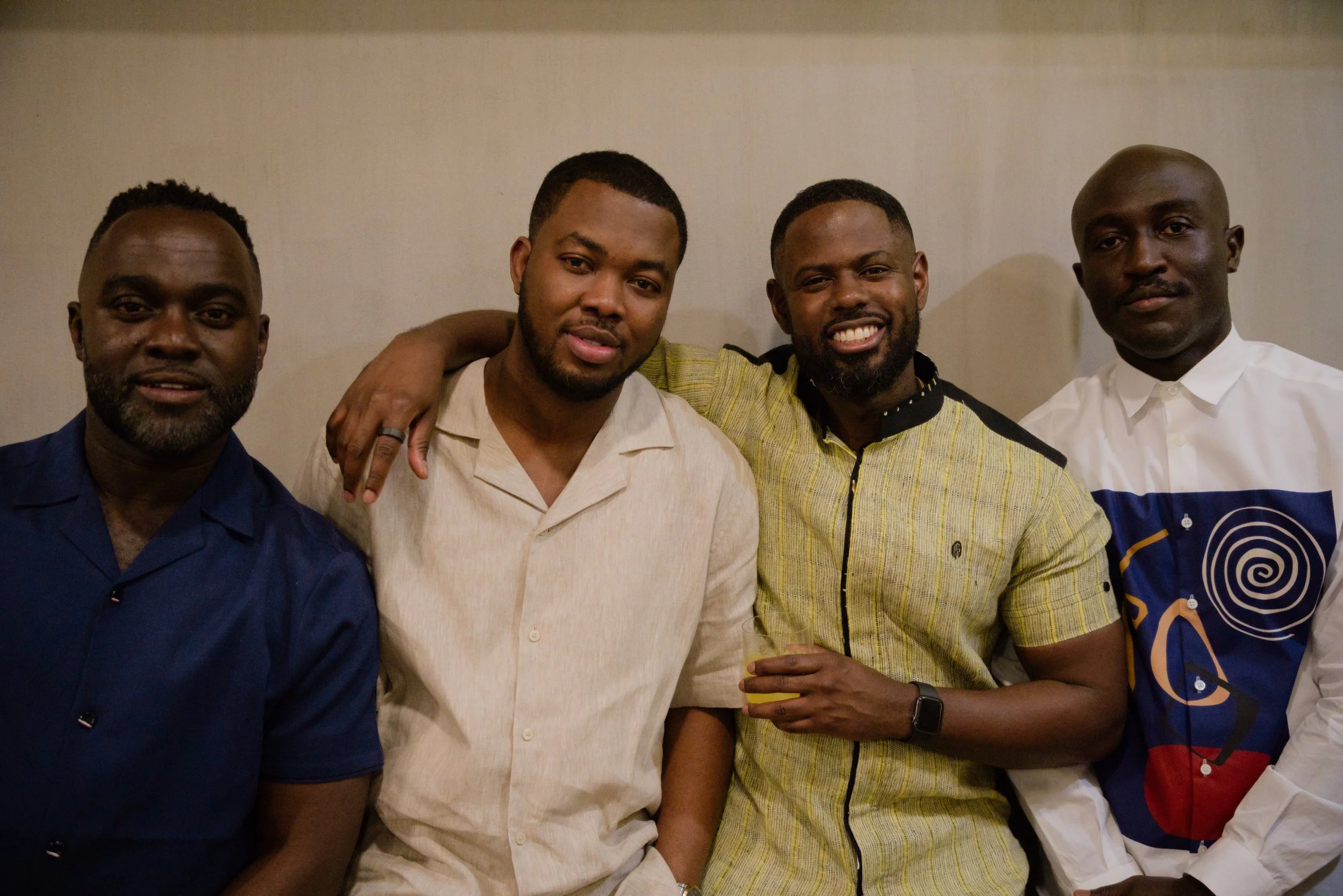 Four men standing together, one with his arm around the others, inside a room with a plain light-colored wall.