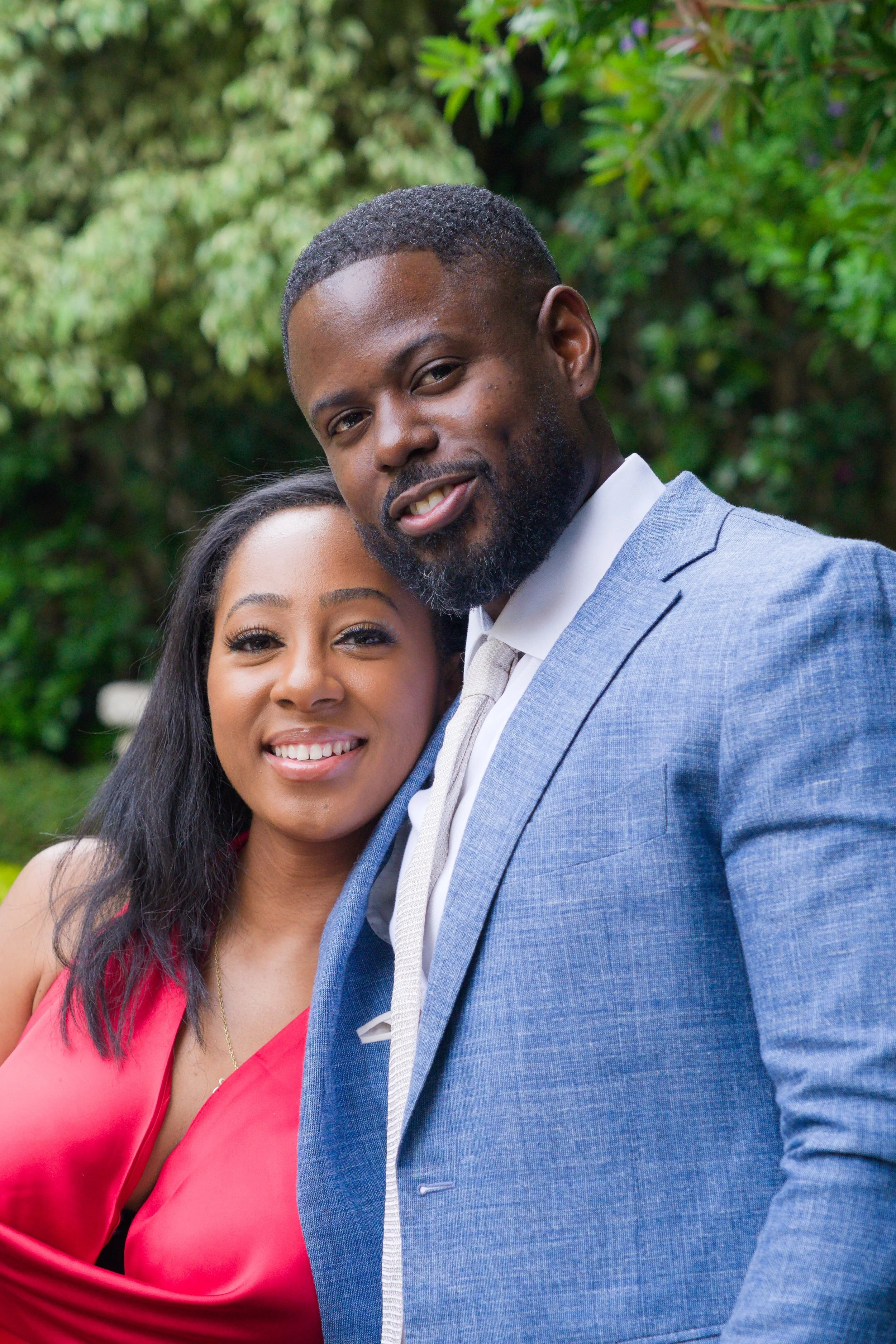 A smiling African-American couple outdoors in front of green foliage, dressed in formal attire. The woman wears a red dress, and the man wears a light blue suit with a white shirt and a tie.
