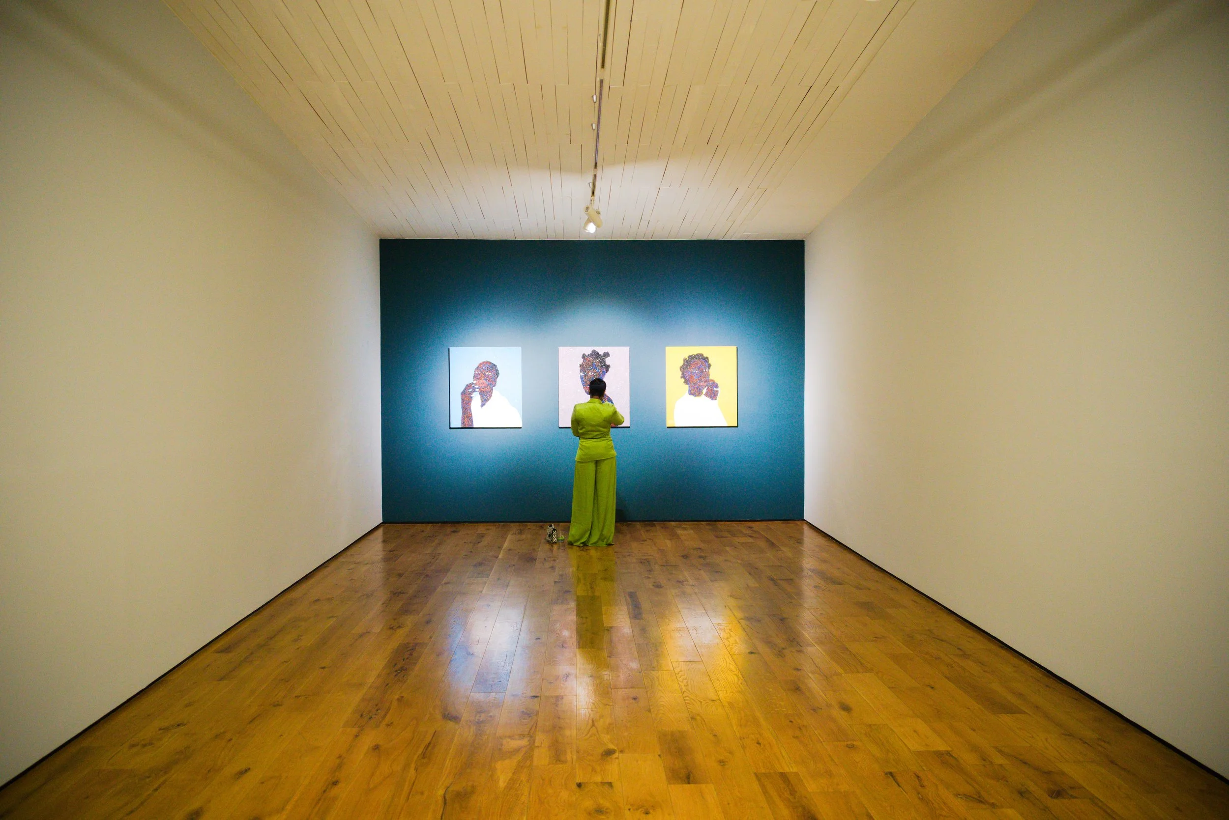 A person in a bright green outfit is standing in an art gallery, facing three contemporary portraits on a blue wall.