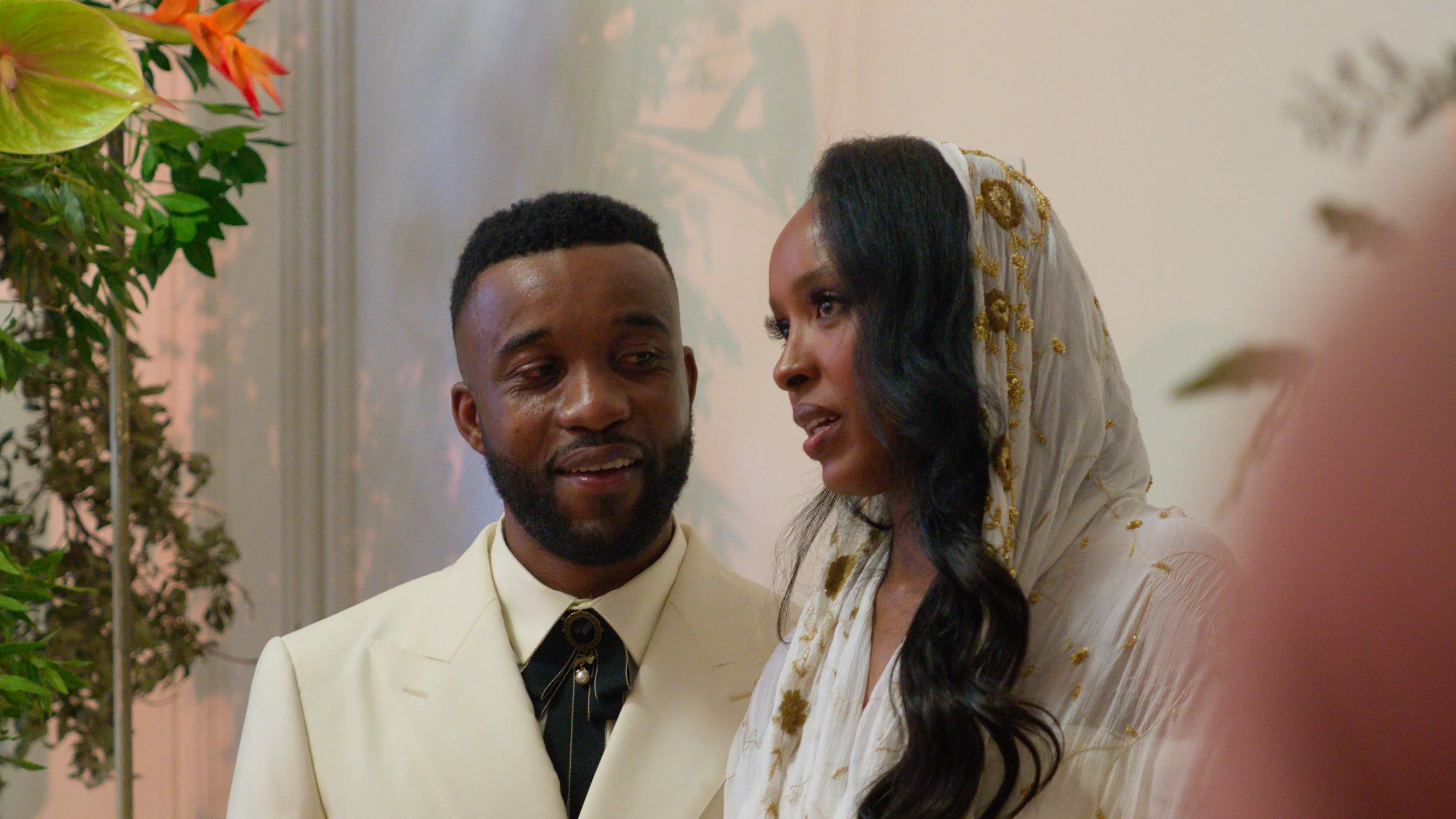 A man and woman are engaged in a close conversation in a well-lit room decorated with plants. The man has a beard and is wearing a cream-colored suit with a black shirt and a bolo tie. The woman has long, dark wavy hair and is wearing a white headsca