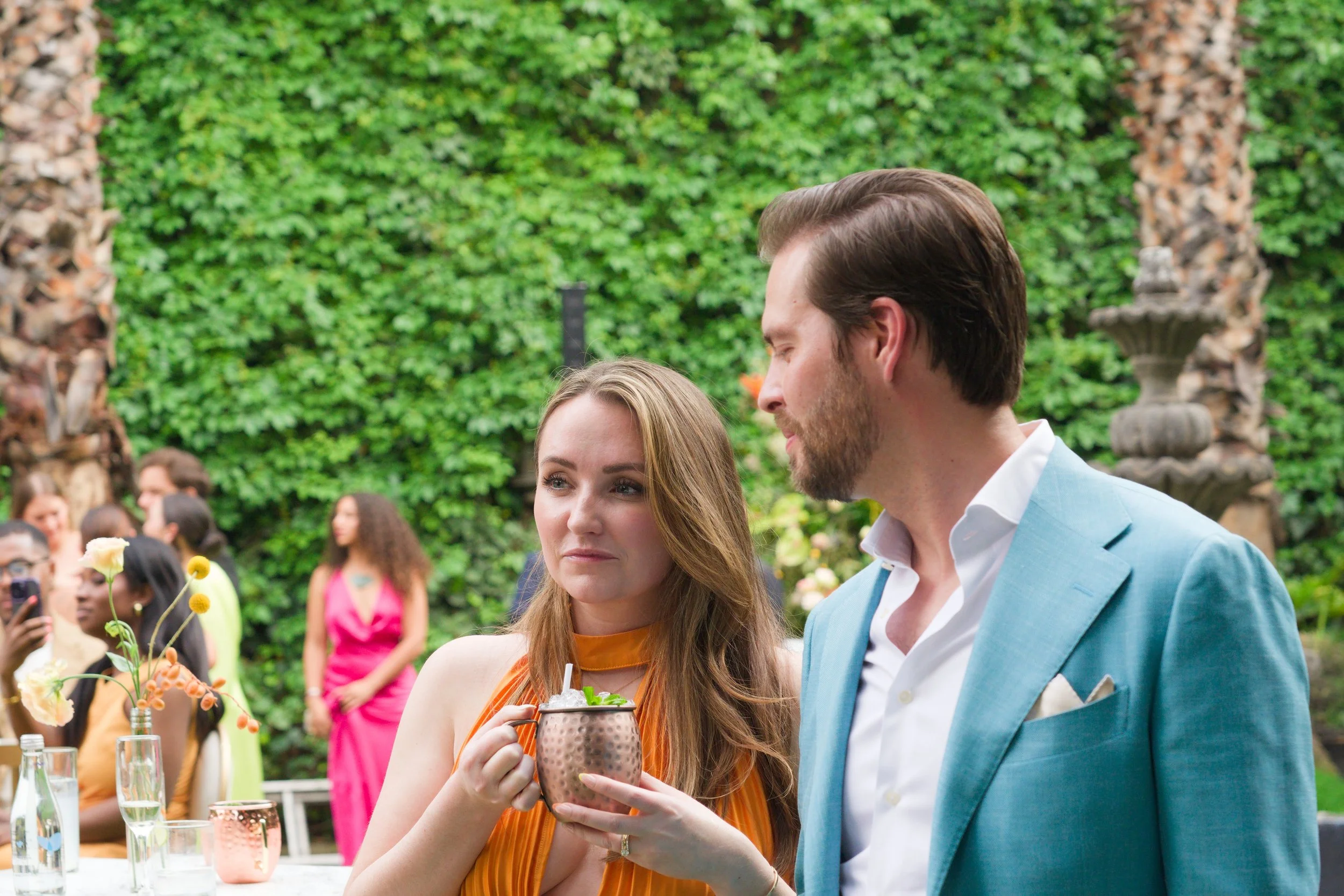 A woman with a drink in a copper mug looking at a man in a blue suit at an outdoor event, with guests in the background and a lush green garden setting.