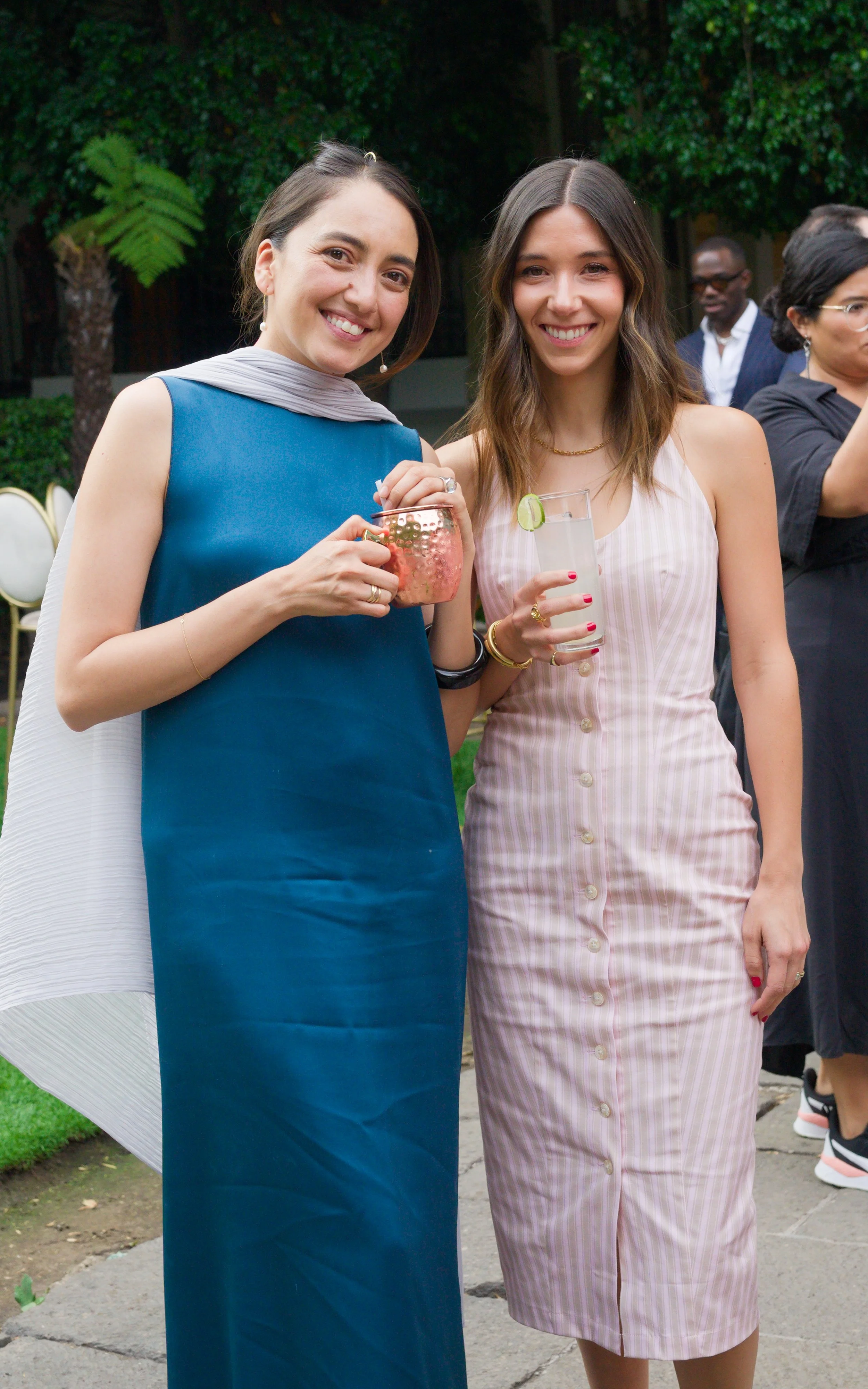 Two women standing outdoors during a social event, smiling and holding drinks, surrounded by a few other people.