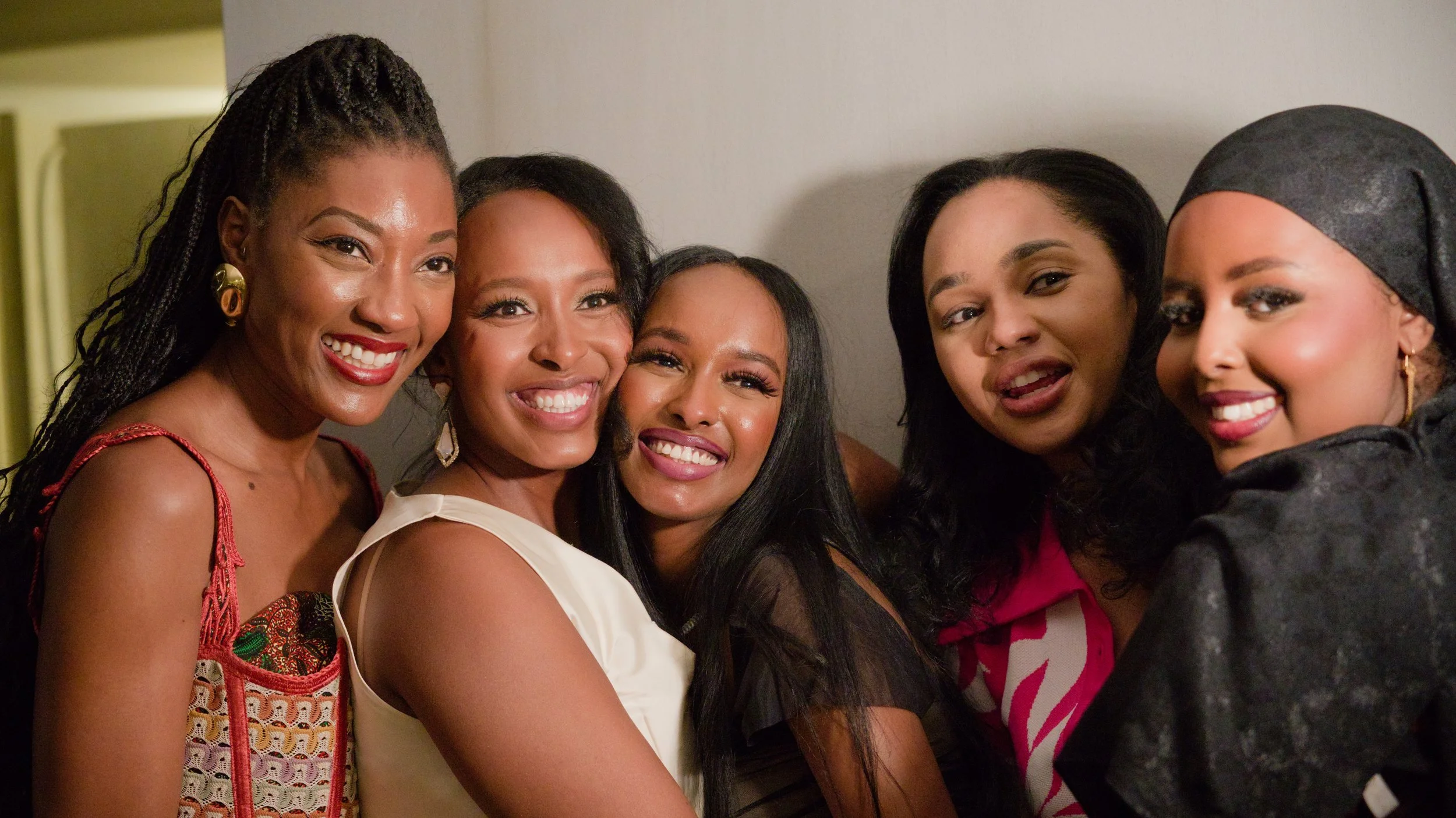 Five women smiling and posing together at a social gathering, wearing stylish outfits and jewelry.