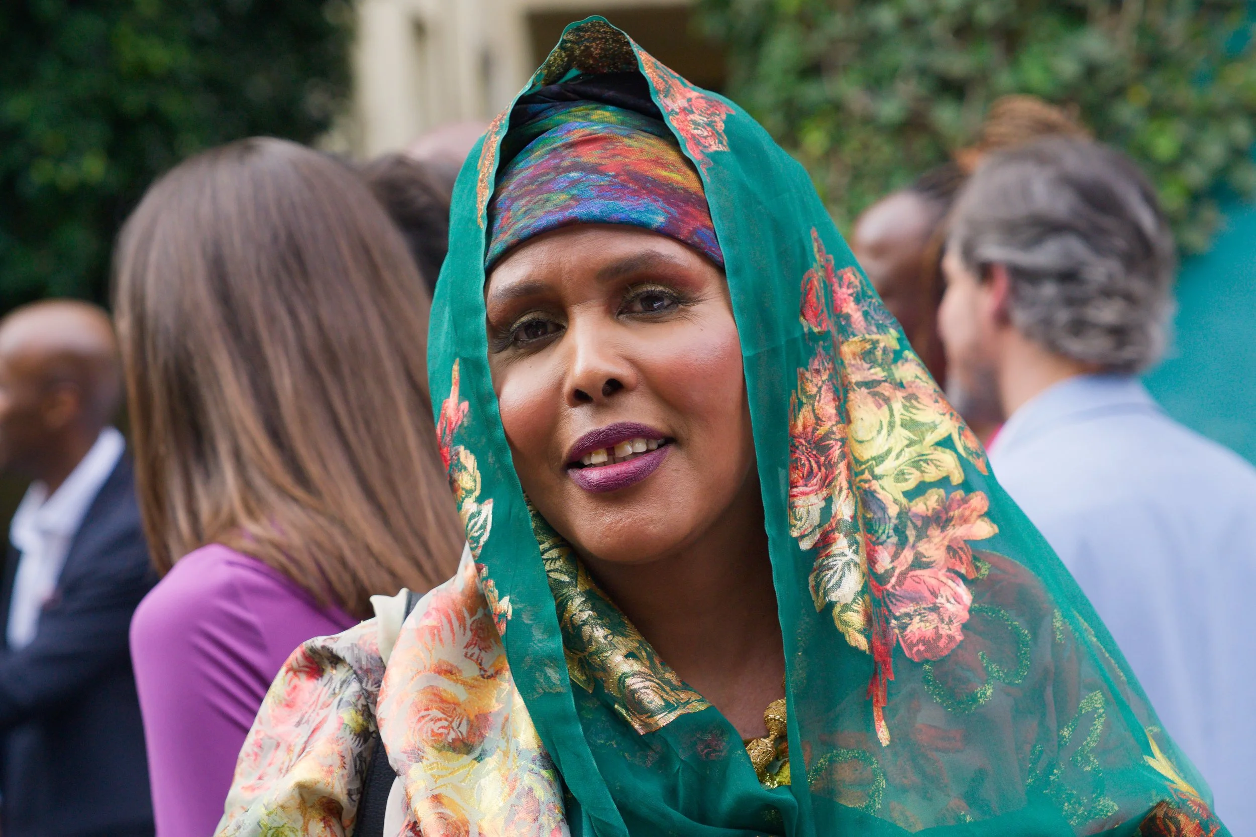 A woman wearing a colorful headscarf and floral-patterned outfit attended an outdoor event among other people.