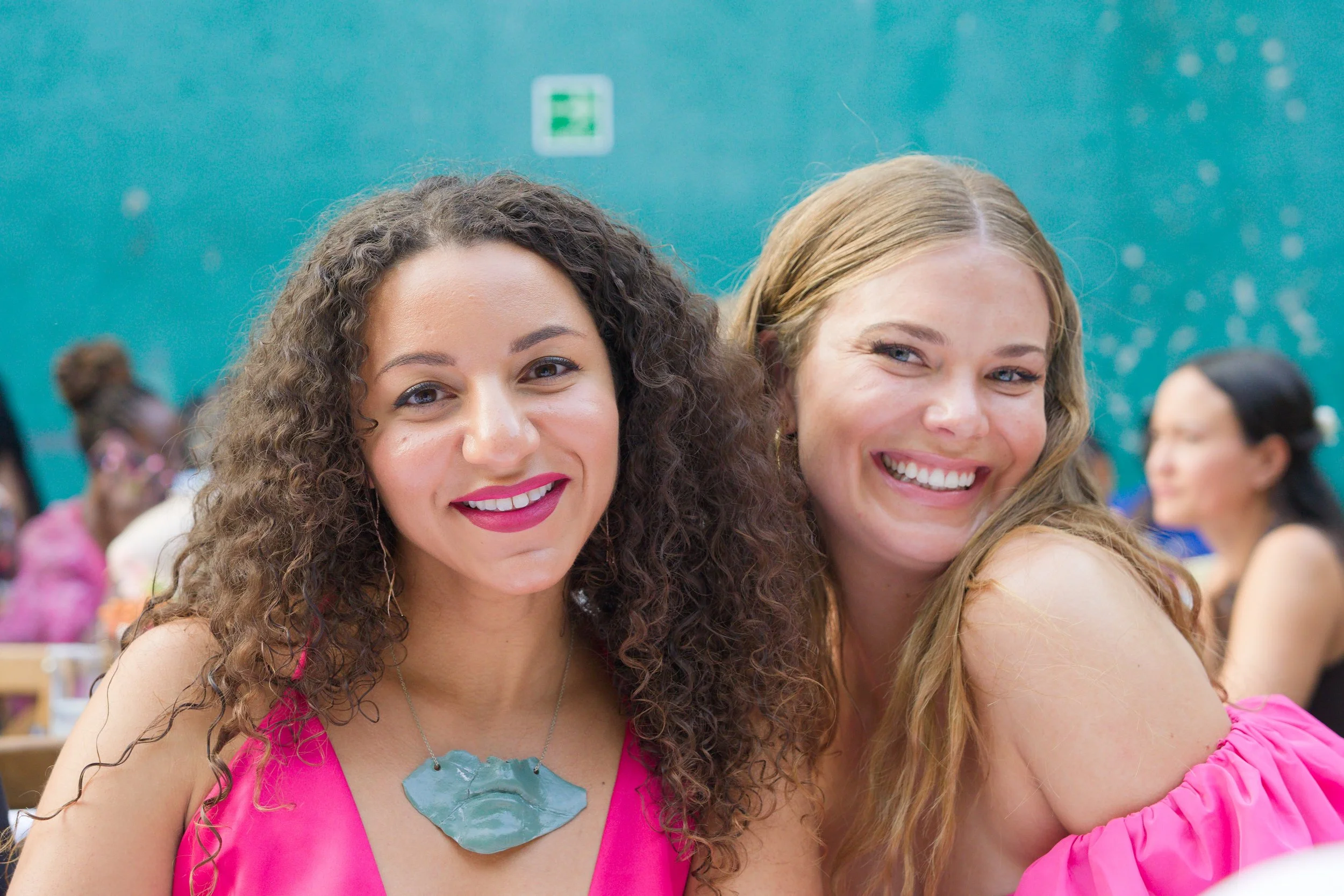 Two smiling women, one with curly hair wearing a pink dress and a necklace, and the other with straight hair wearing a pink off-shoulder dress, sitting at a table in a room with a teal-colored wall and other people in the background.