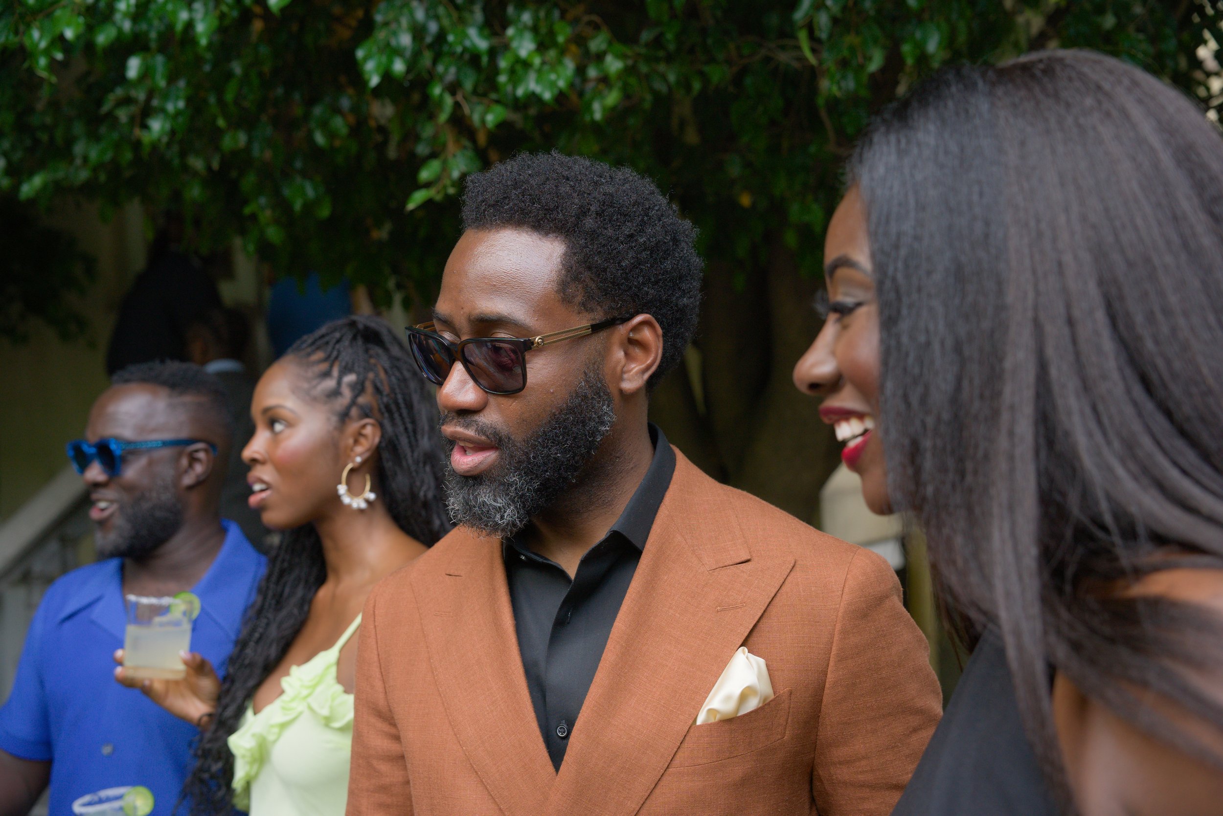 Group of people at a social gathering outdoors, dressed in stylish clothes, with trees in the background.