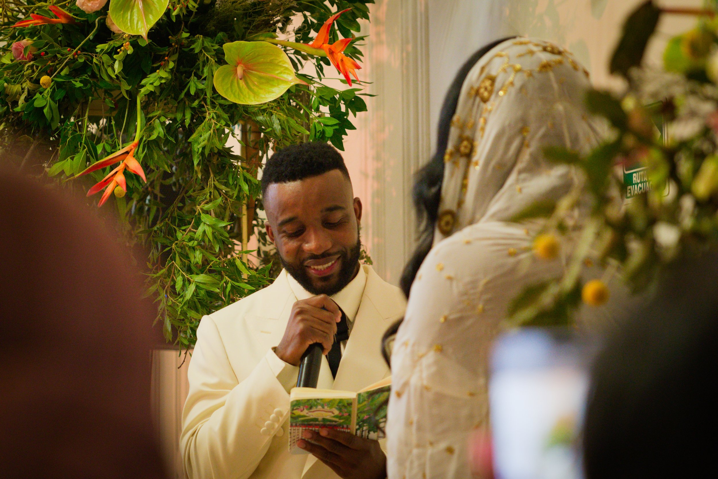 A man in a white suit holding a microphone and reading from a book during a ceremony, with a woman in a traditional embroidered dress facing him, surrounded by green plants and flowers.