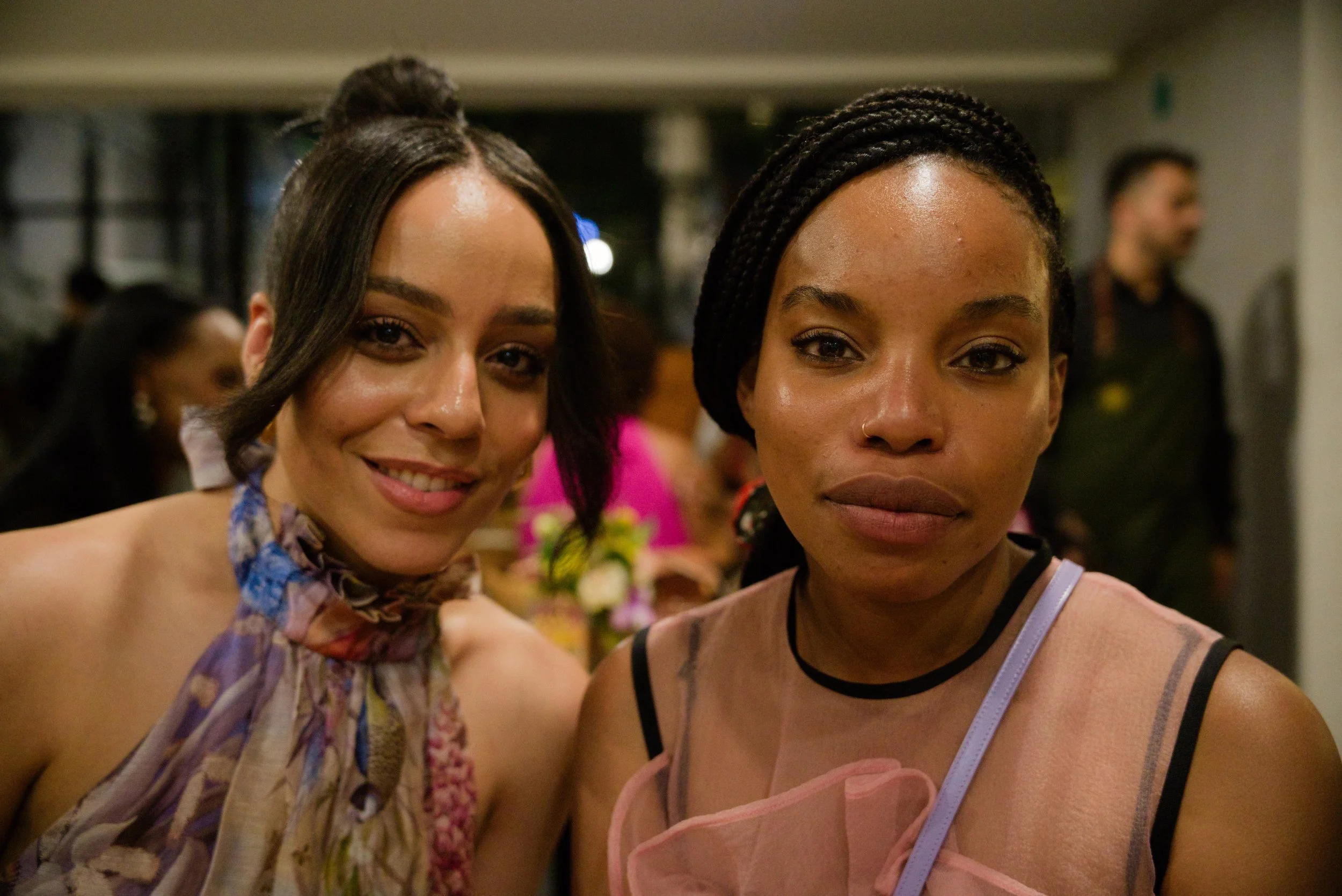 Two women posing for a photo at an indoor event, with a blurred background of other people at a gathering.