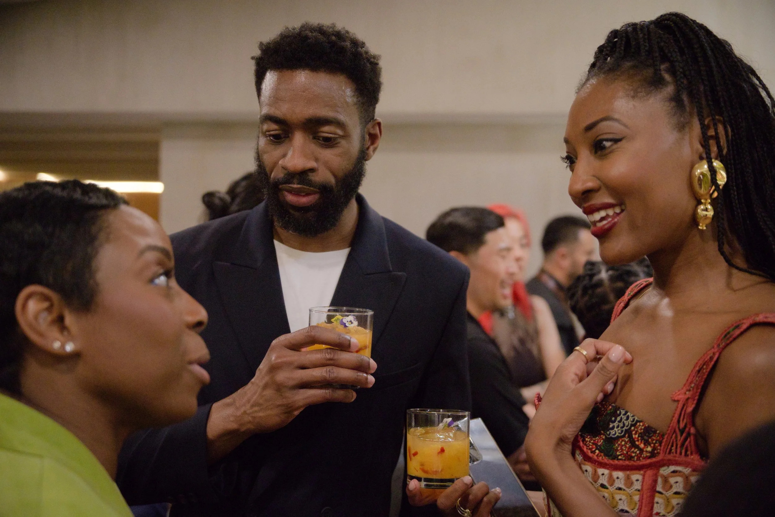 People socializing at a party, three women and one man in the foreground, holding drinks, with others in the background.