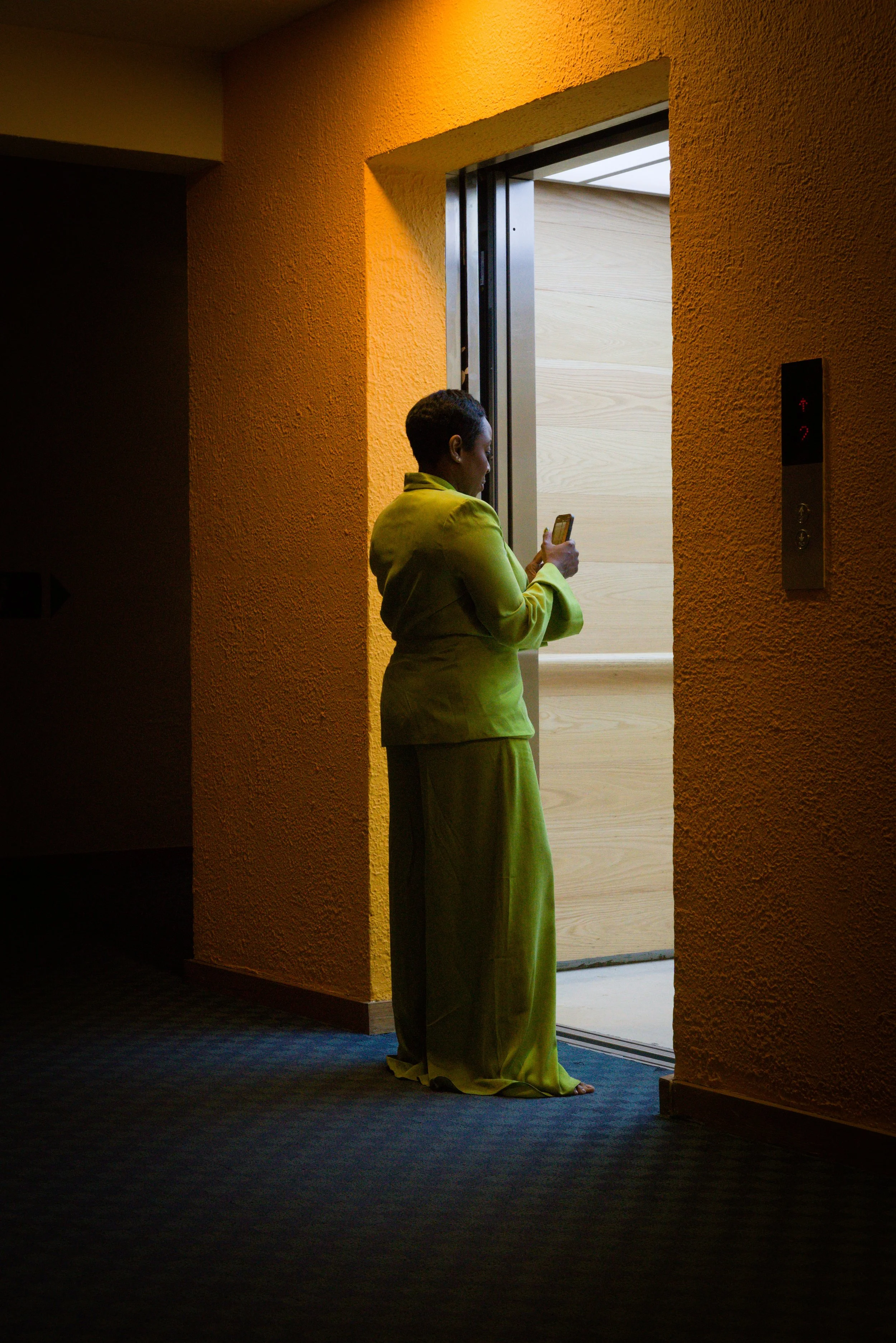A woman in a bright green suit standing in front of an elevator, looking at her phone. The elevator doors are open, revealing a light wood interior.