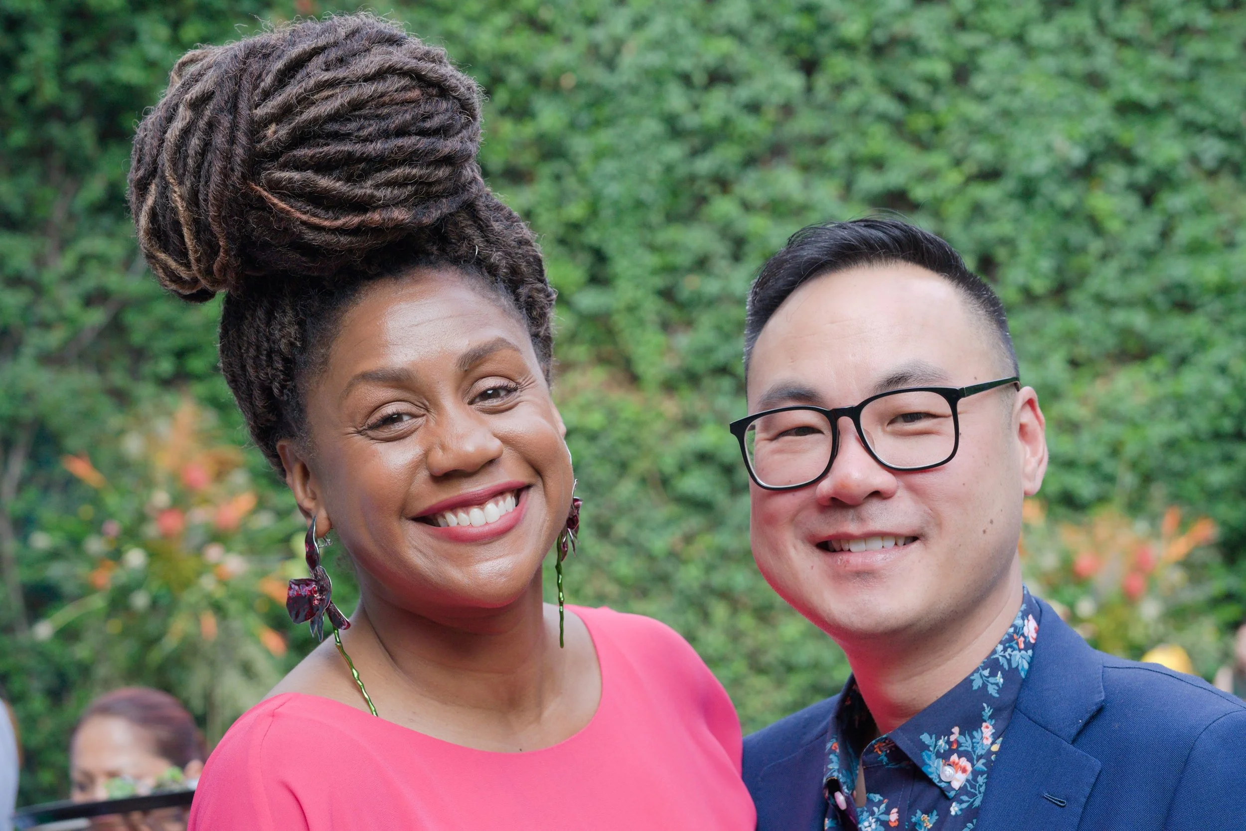 Smiling woman with large braided hairstyle wearing a pink dress standing next to a smiling man with glasses in a blue suit and floral shirt, outdoors with green trees in the background.