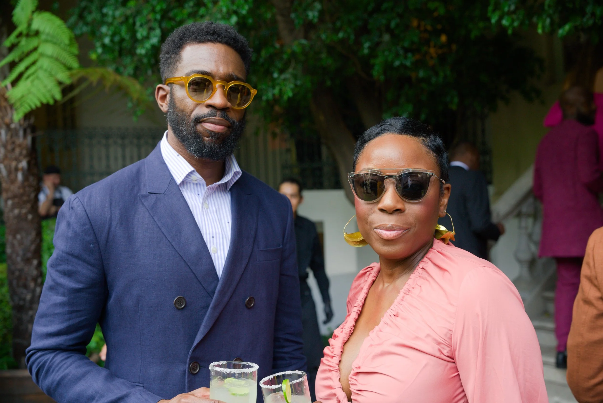 A man and woman at an outdoor event, both wearing sunglasses, holding drinks with lime slices, dressed in semi-formal attire, with a background of other guests and greenery.