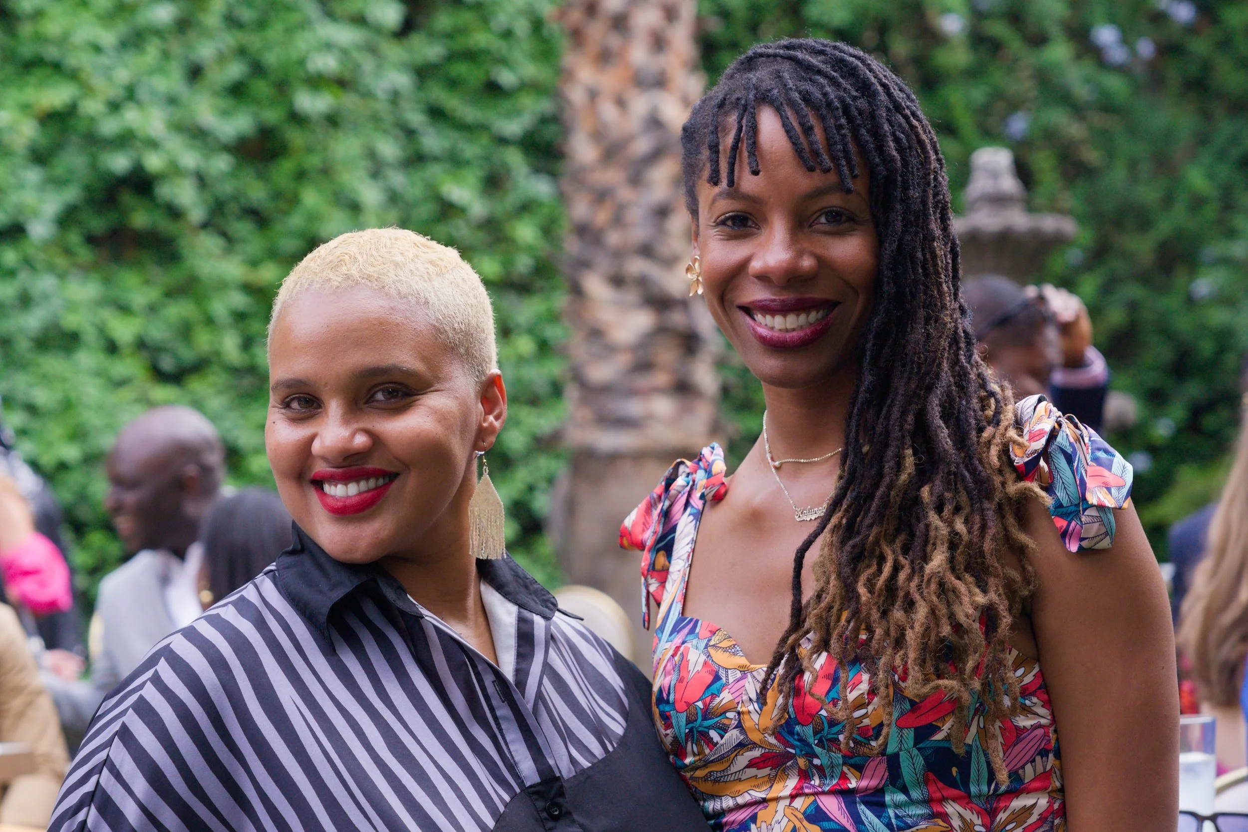 Two women smiling outdoors at a gathering or celebration, with greenery and trees in the background.