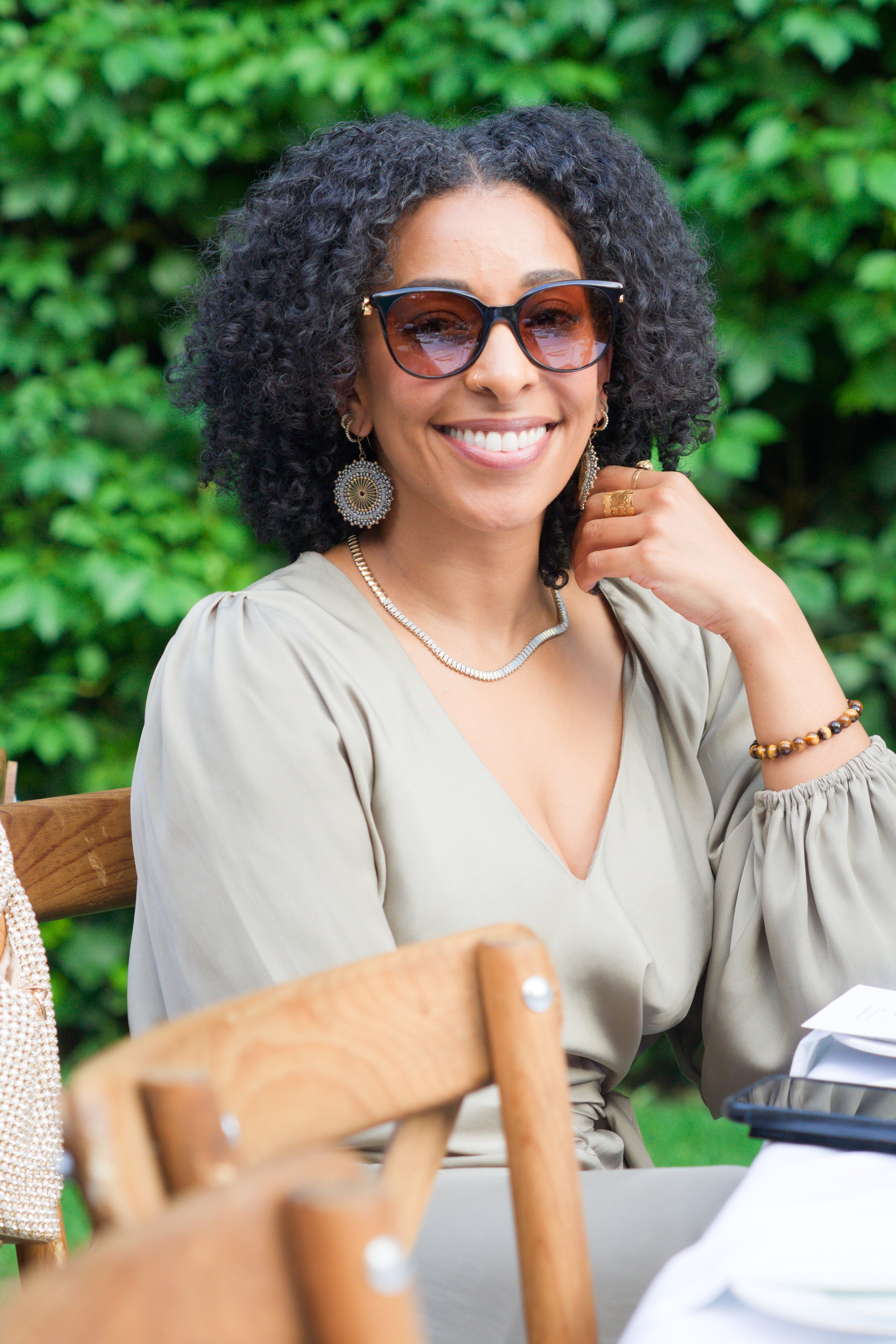 A woman with curly black hair wearing sunglasses, jewelry, and a beige top, sitting outdoors with green foliage in the background, smiling at the camera.