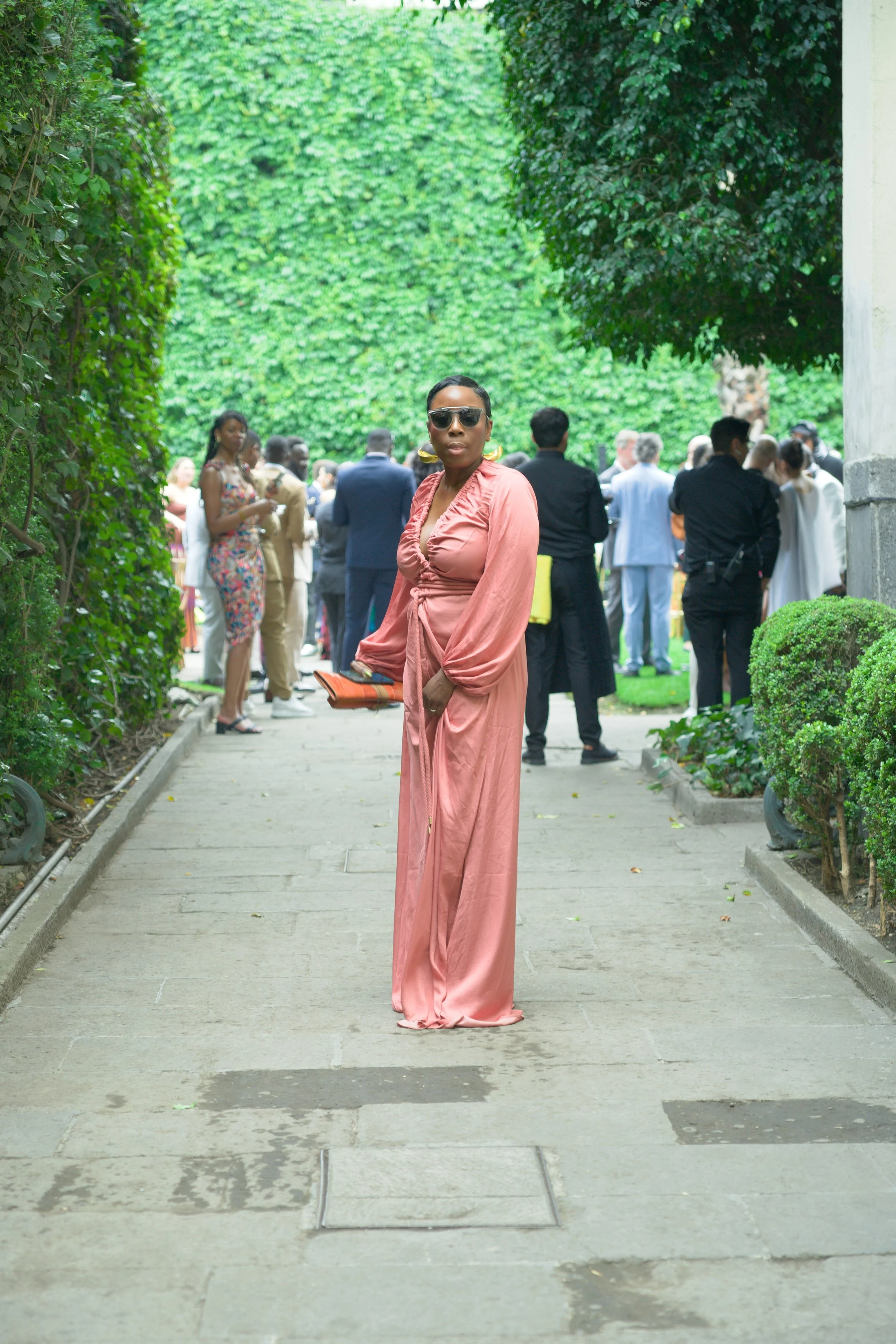 A woman in a long pink dress and sunglasses standing on a walkway outdoors among many people dressed formally at an event.